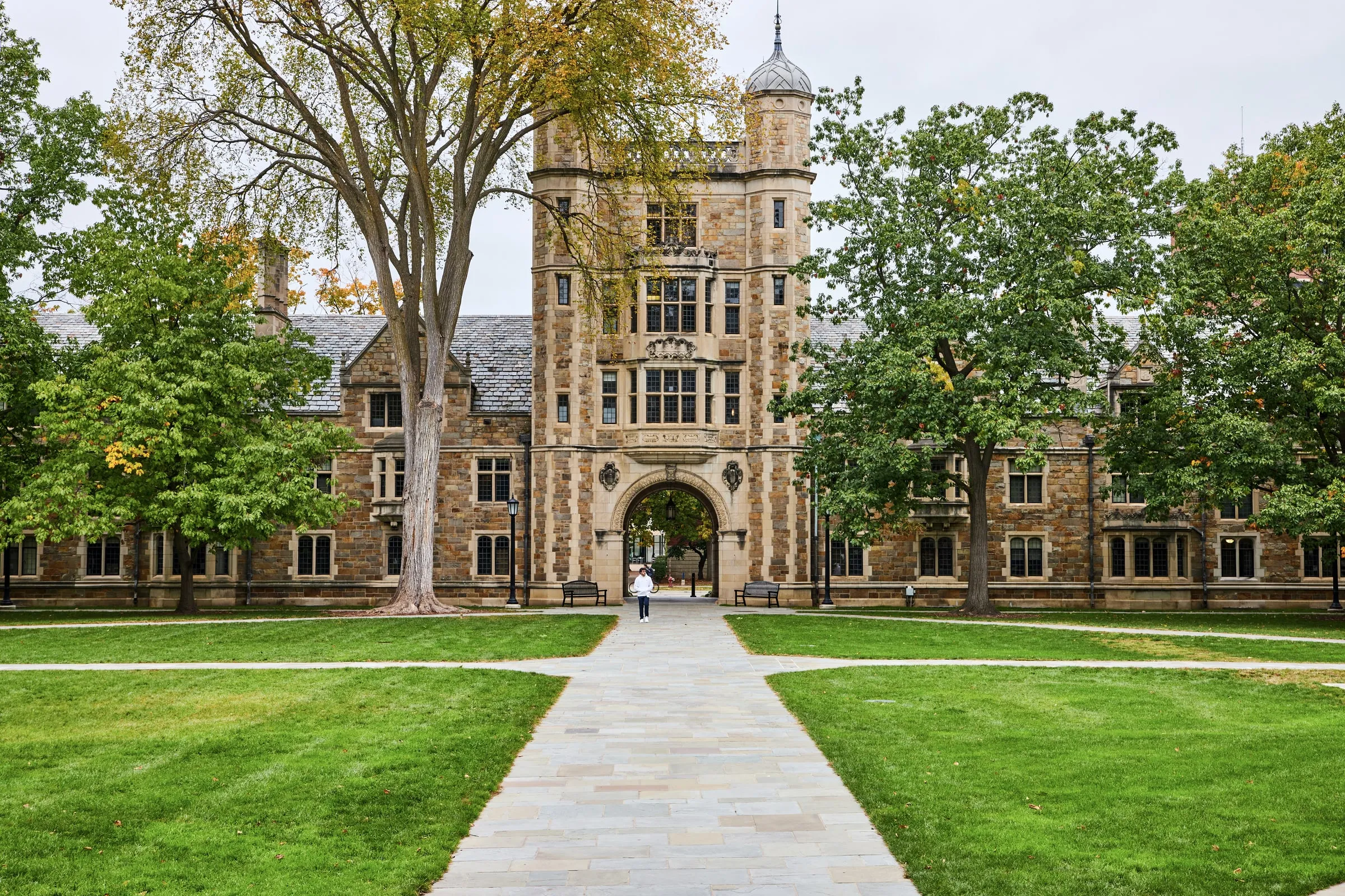 Stone university building with an arched passageway and turret, viewed across a lawn and paved walkway lined with trees