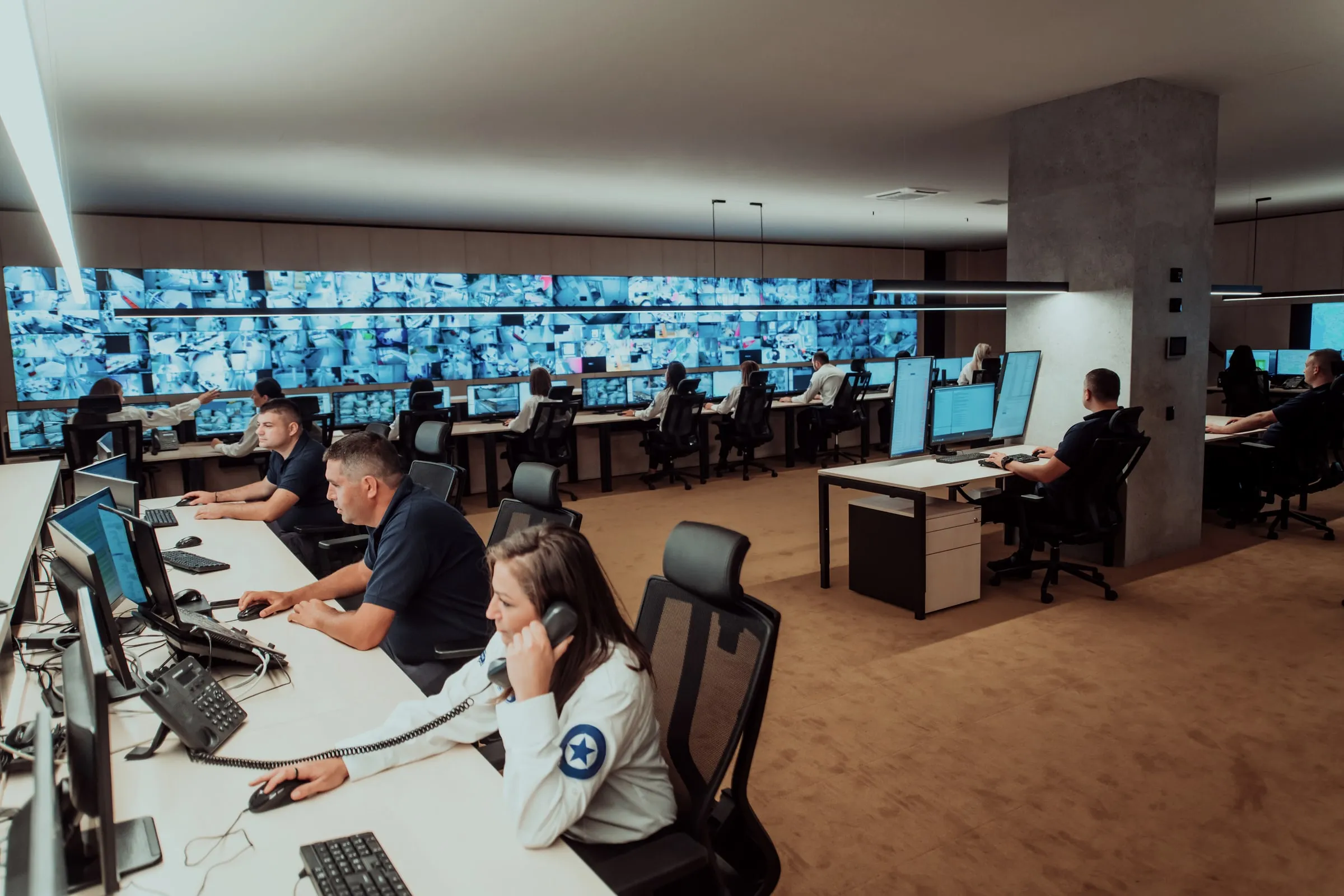 Open security monitoring center with staff at rows of desks below a large wall of live camera feeds, one officer speaking on a phone in the foreground