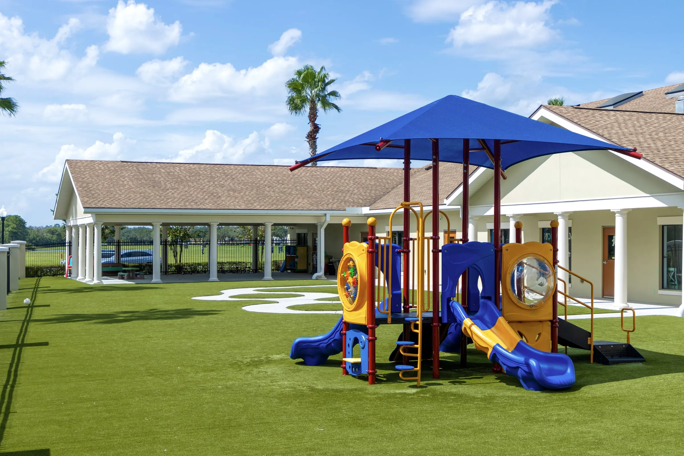 Children's playground with a blue shade canopy over a play structure on artificial turf, beside a single-story building with a covered walkway