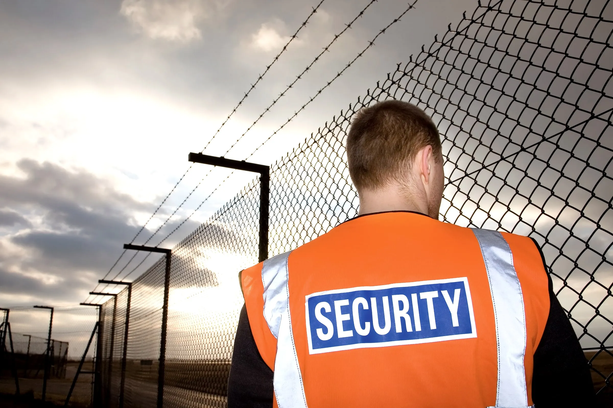 Security officer in an orange vest marked SECURITY viewed from behind, facing a chain-link fence topped with barbed wire under a cloudy sky