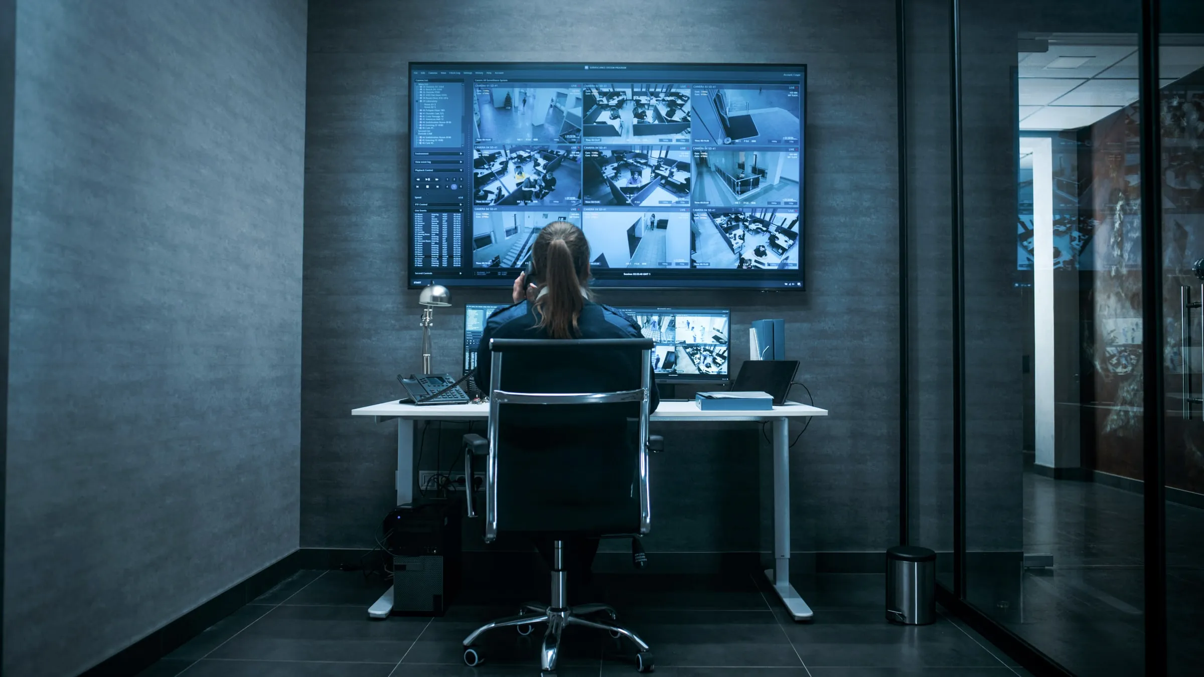 Security staff member seated at a desk speaking on a phone while viewing a wall of monitors showing live camera feeds from several rooms