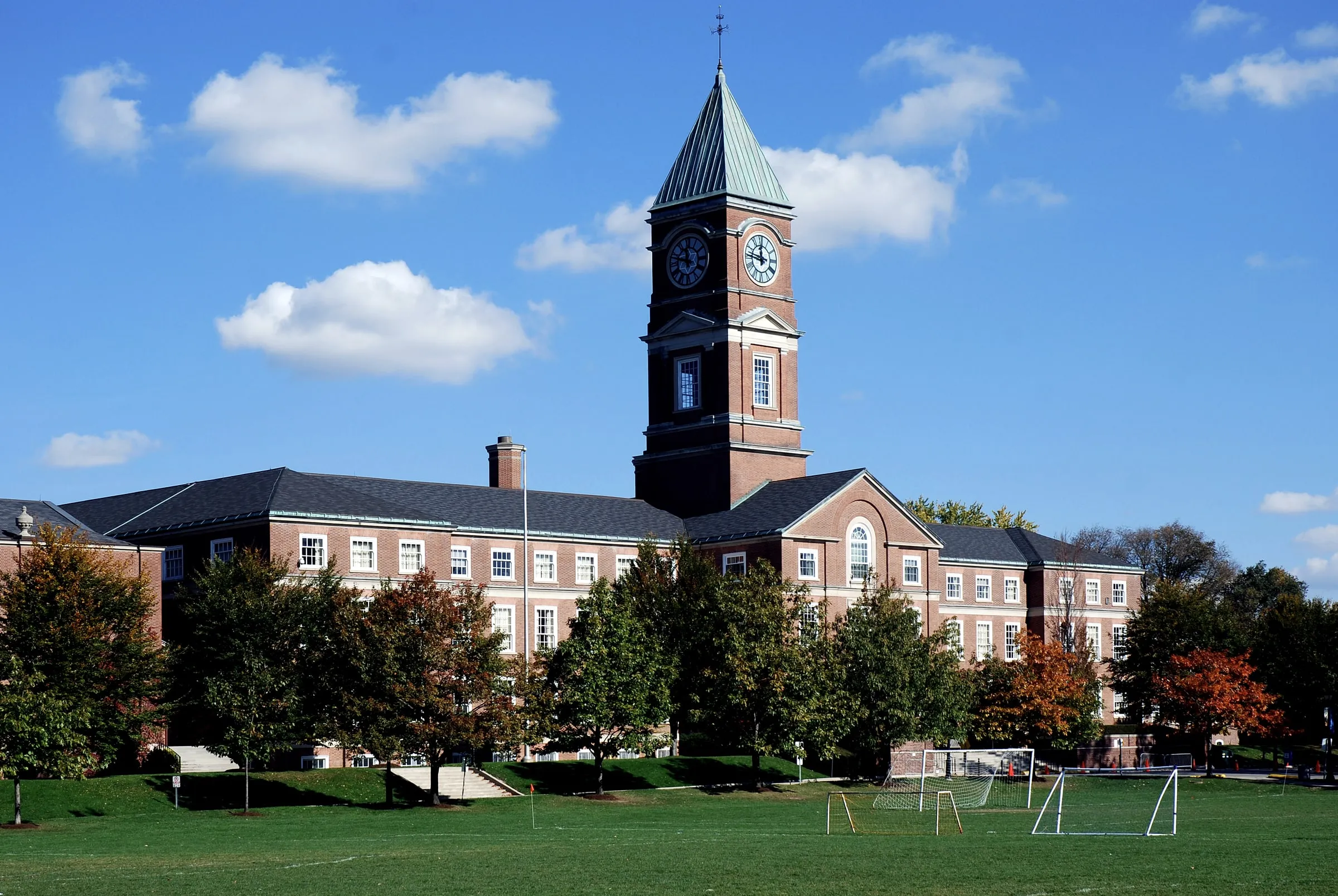 Brick school building with a tall clock tower, columned entrance and a playing field with soccer goals, framed by trees turning autumn color