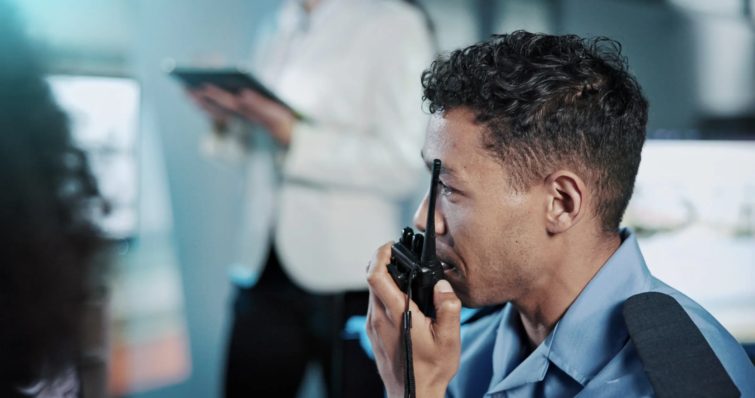 Close-up profile of a man in a collared shirt speaking into a handheld radio, with a person holding a clipboard blurred in the background