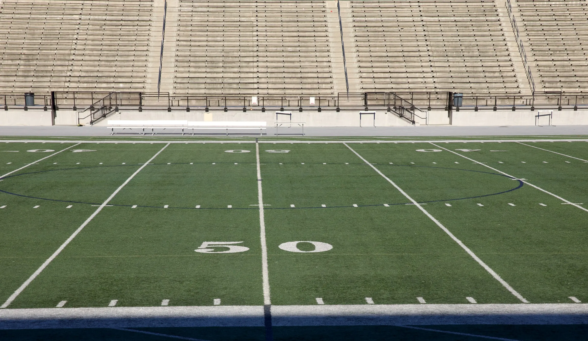 Empty football stadium bleachers above an artificial turf field marked at the 50-yard line