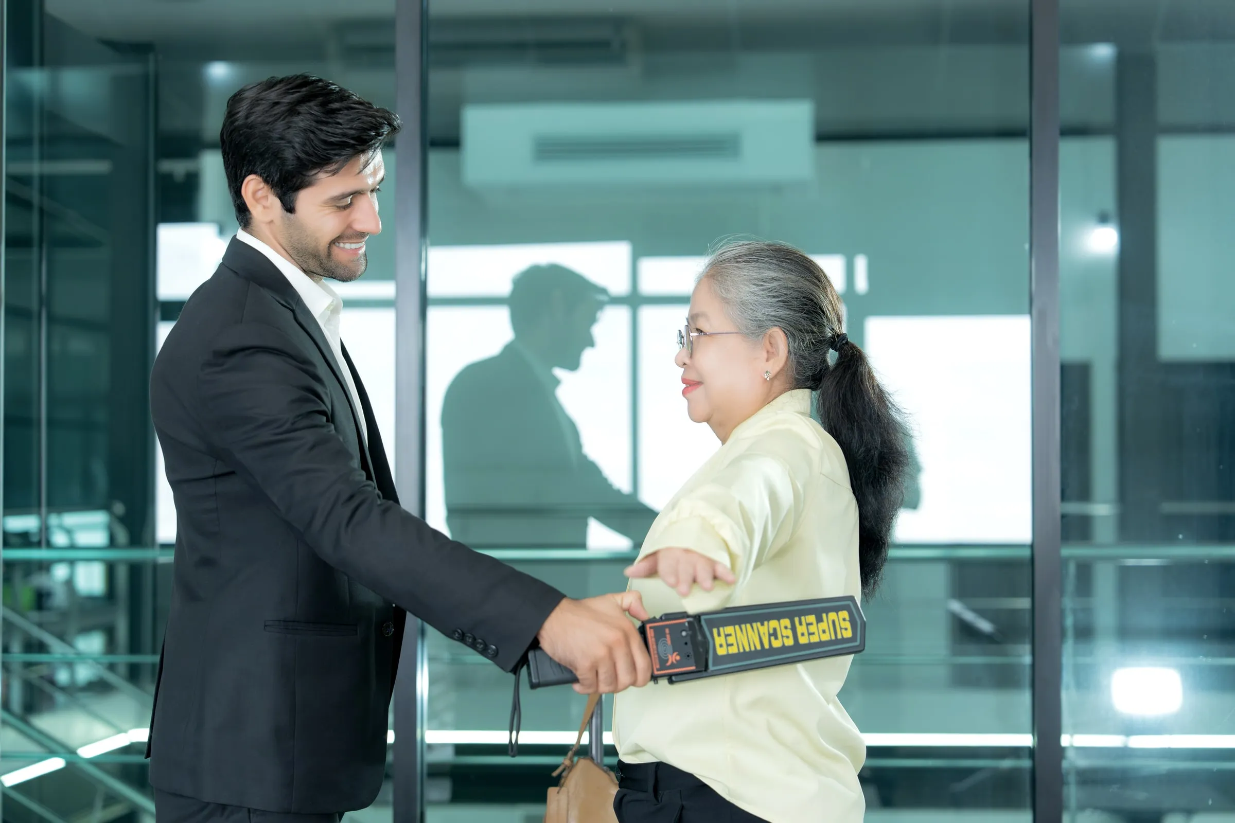 Security officer in a dark suit screening an older visitor with a handheld metal detector wand at a glass entrance