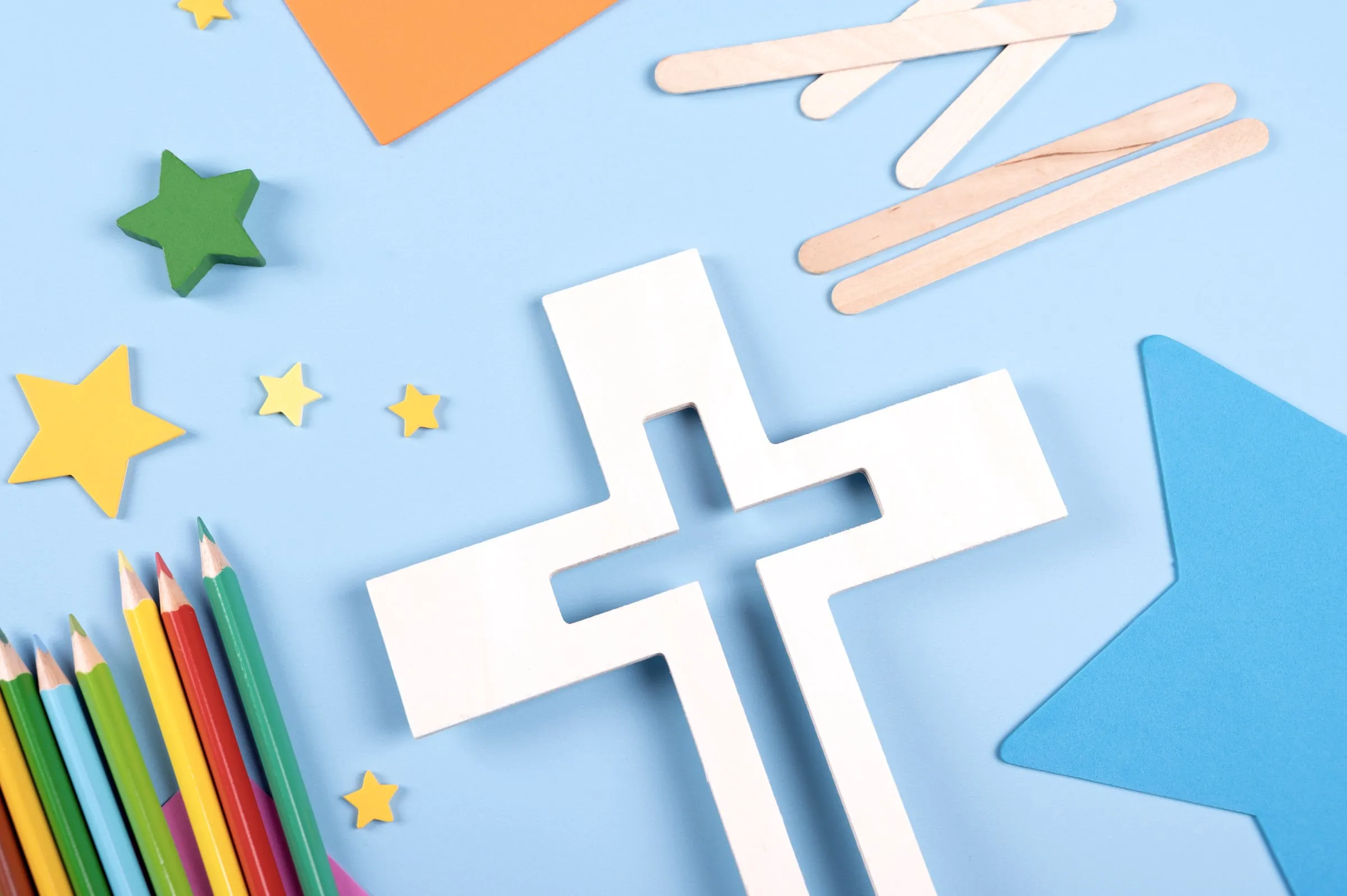 White paper cross, colored pencils and paper stars laid out on a blue background