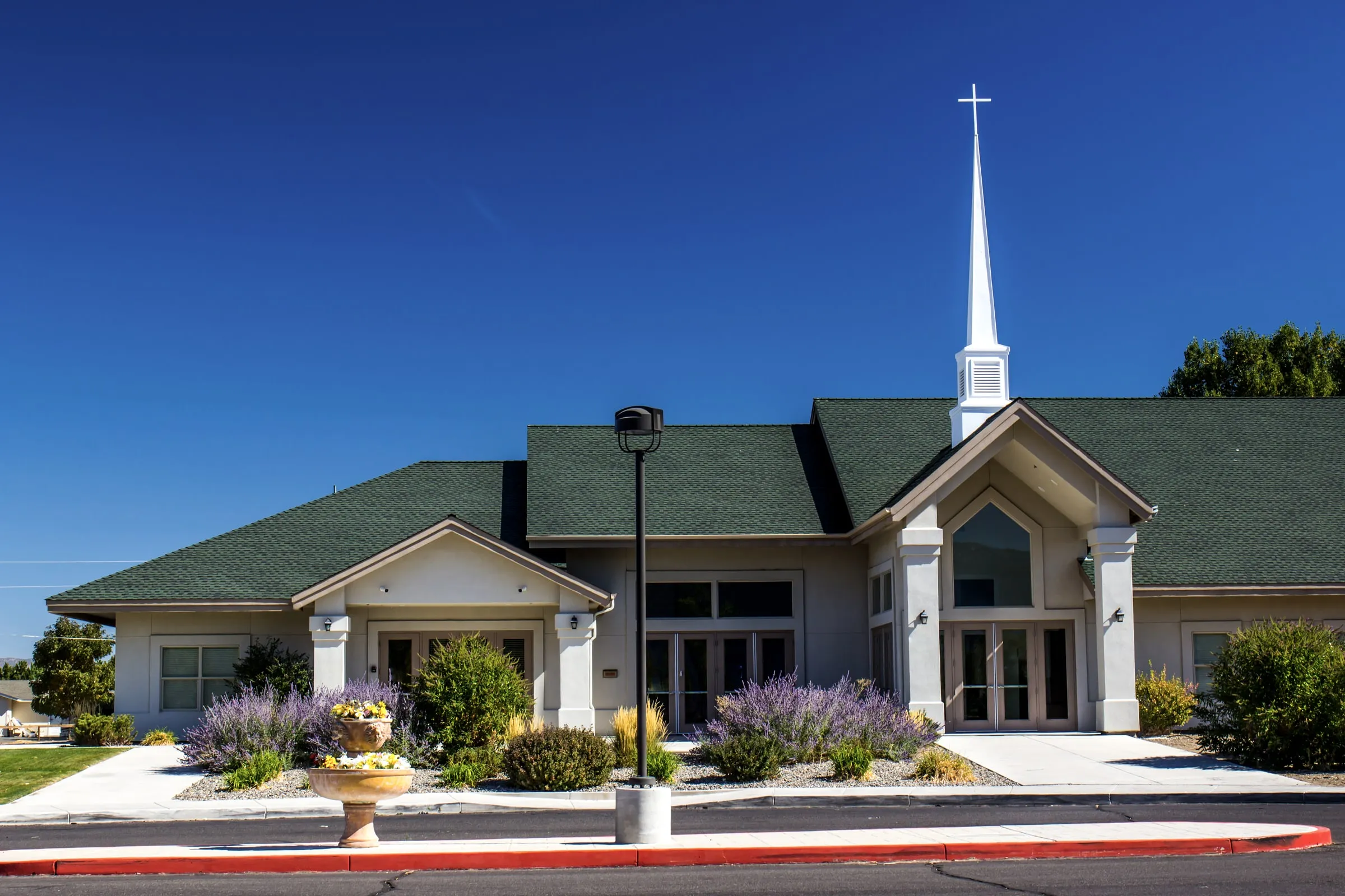 Small church with a white steeple, green roof and purple flowers in front under a clear sky