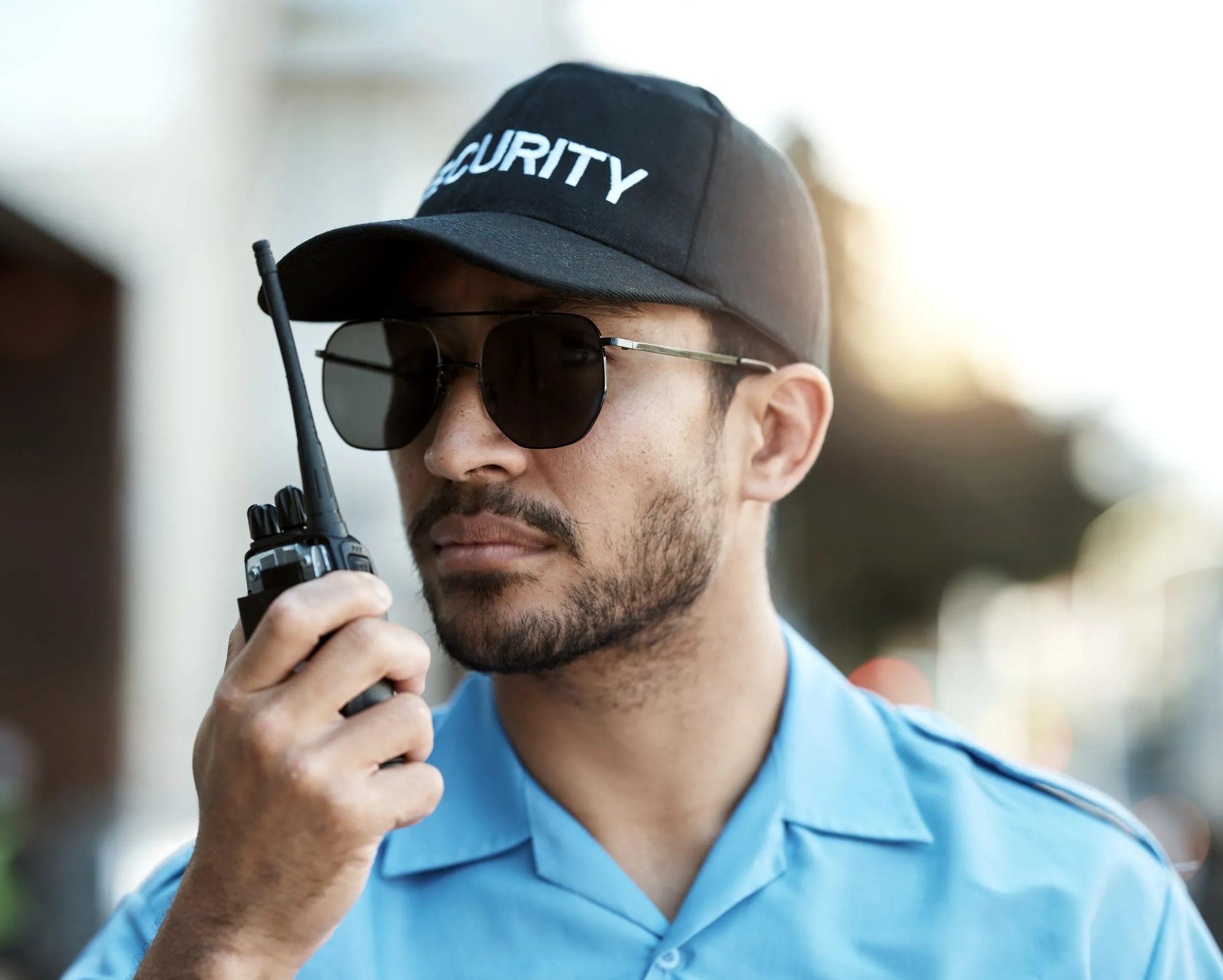 Security officer in a black SECURITY cap and sunglasses speaking into a radio on a bright street