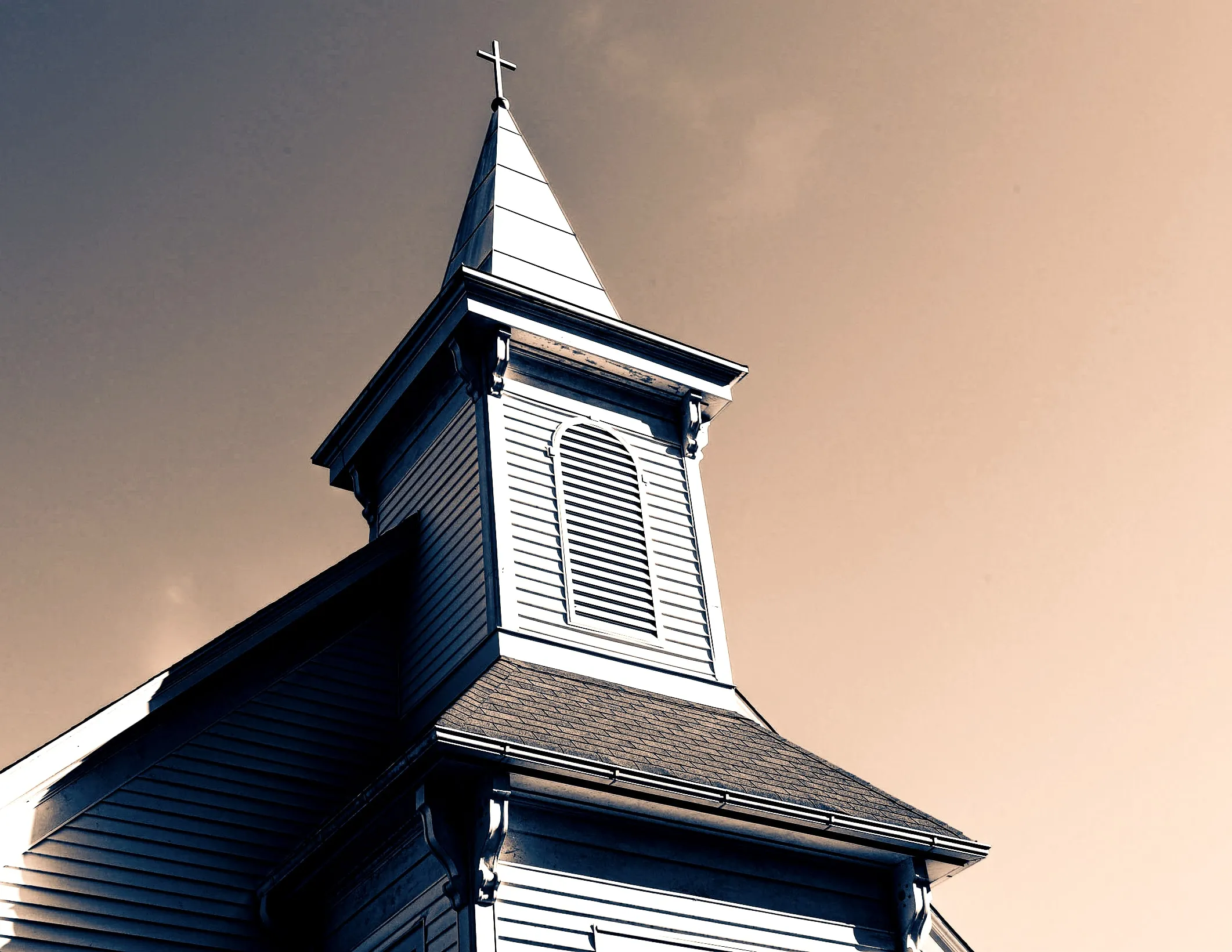 Church steeple with louvered belfry and a cross against a hazy golden sky