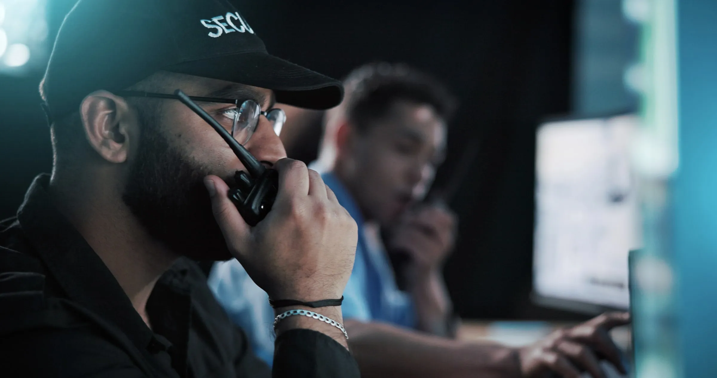 Security officer in a black cap speaking into a radio in a dim control room beside a colleague at a monitor