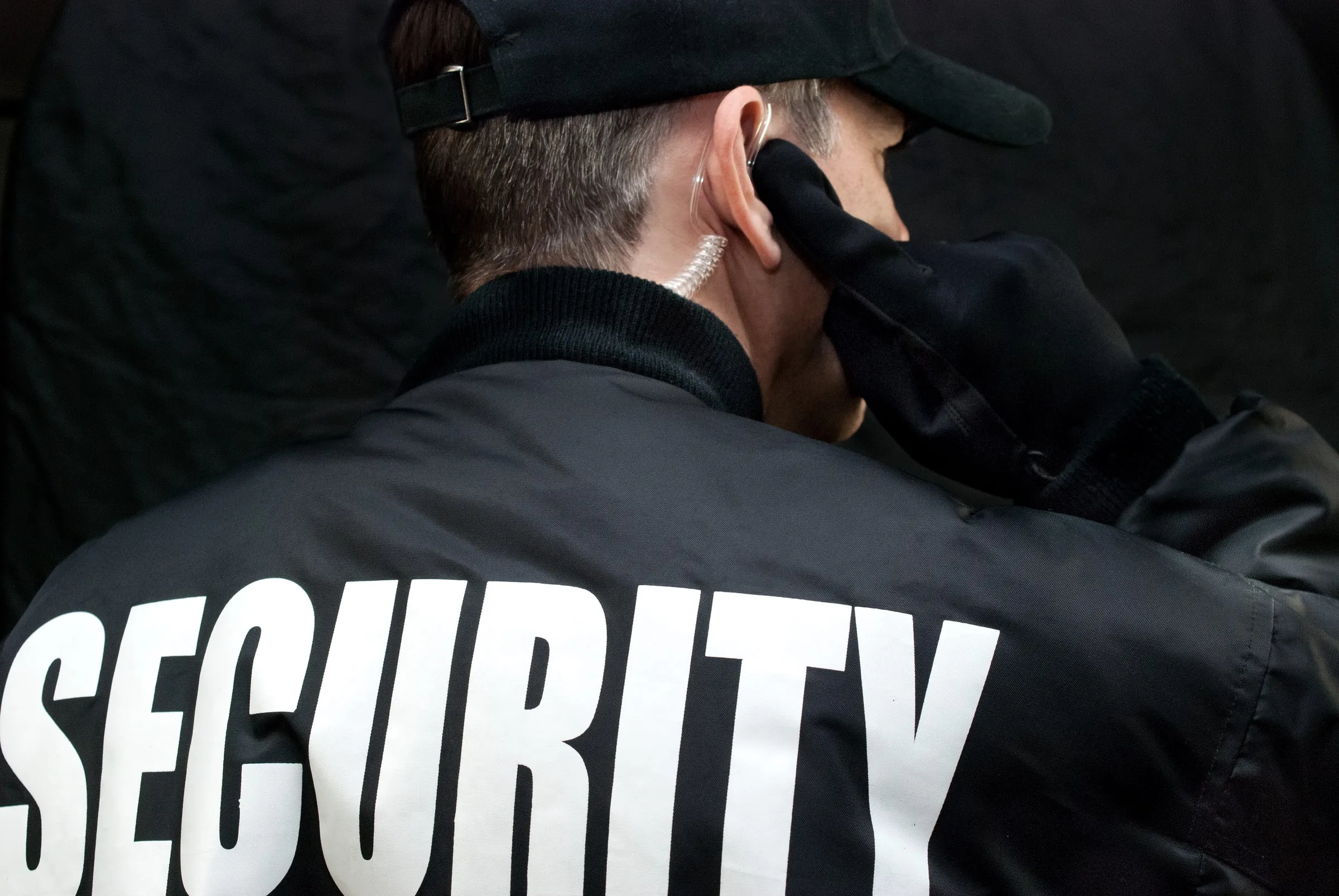 Back of a security officer's black jacket with SECURITY lettering, hand at an earpiece
