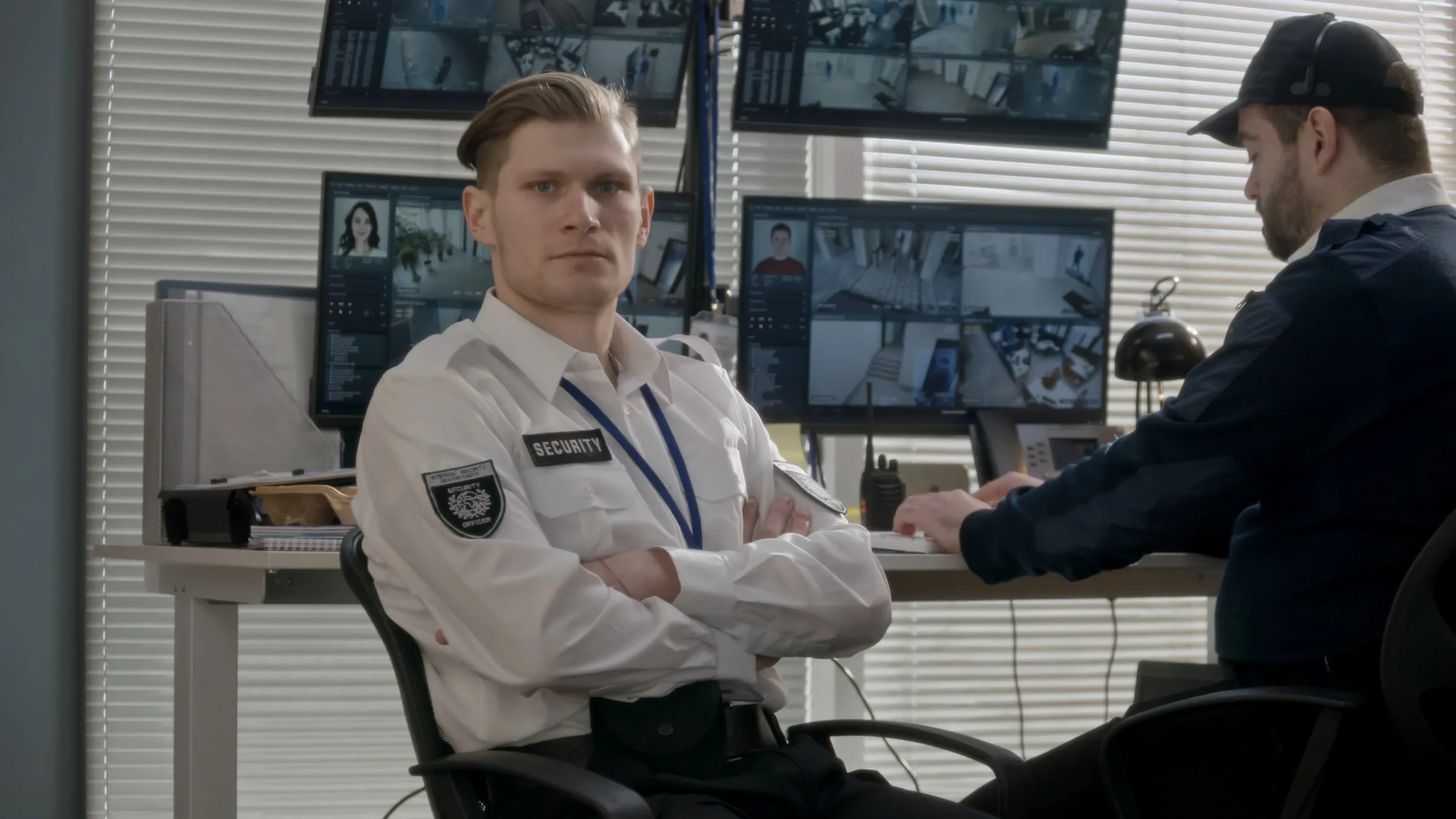 Security officer in a white shirt seated at a desk with monitors, looking at the camera