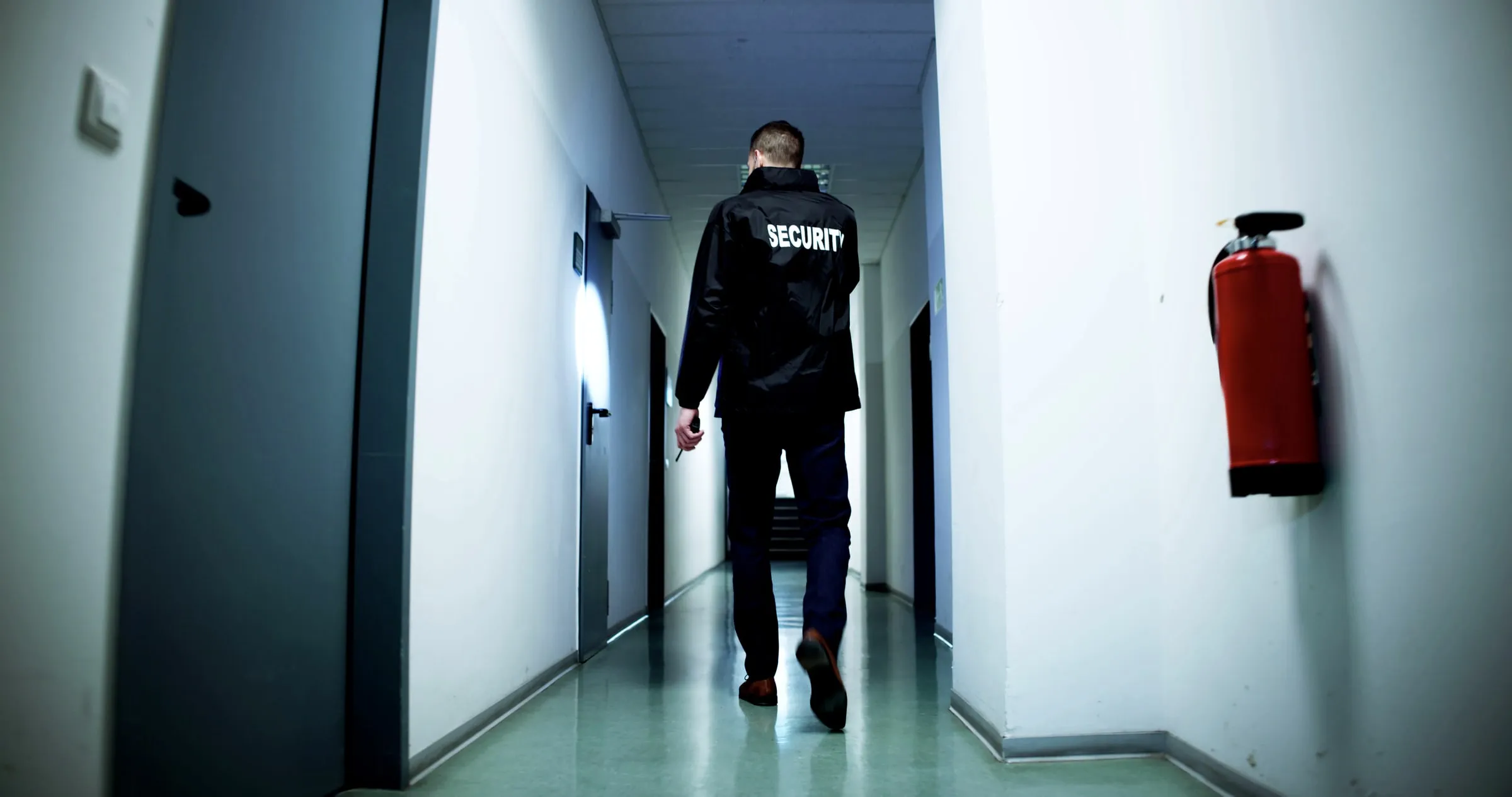 Officer seen from behind wearing a jacket marked Security, walking down a corridor past a wall-mounted fire extinguisher and closed doors