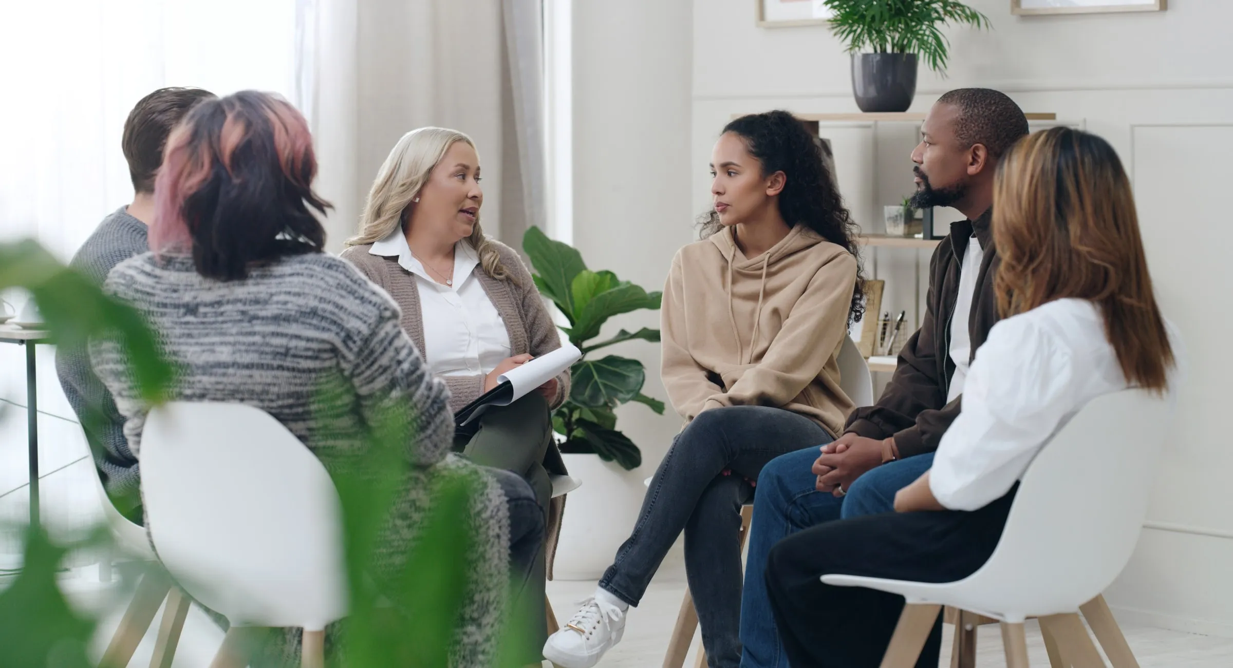Six adults seated in chairs arranged in a loose circle in a bright, plant-filled room, in conversation with a facilitator holding a notepad