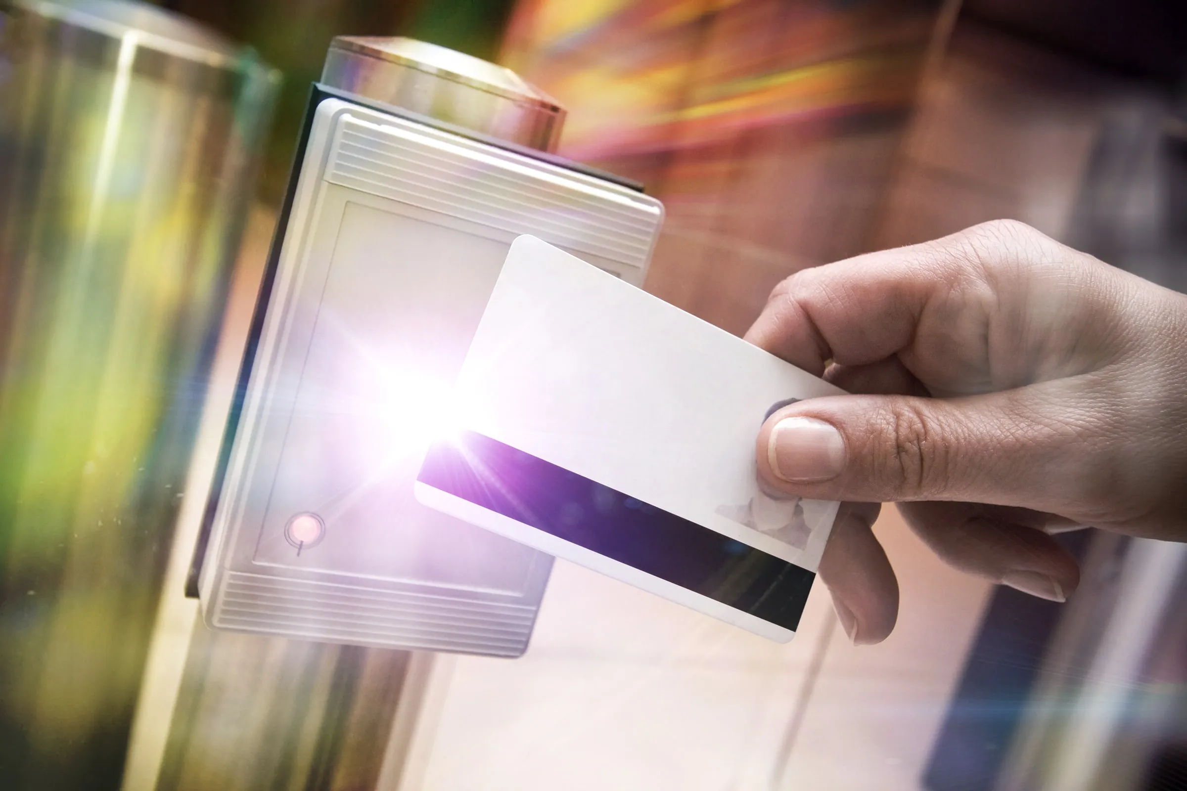 Hand holding a white access card in front of a glowing card-reader mounted on a post, with a blurred, colorful background