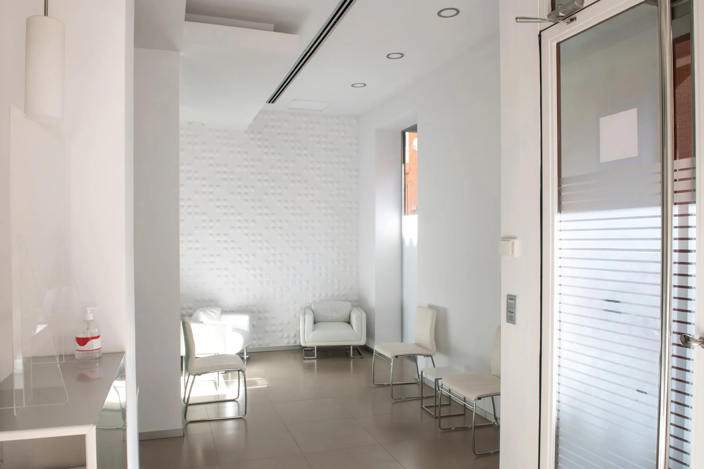 White minimalist waiting room with leather armchairs and chairs, a textured tiled accent wall, and a hand sanitizer station on a counter
