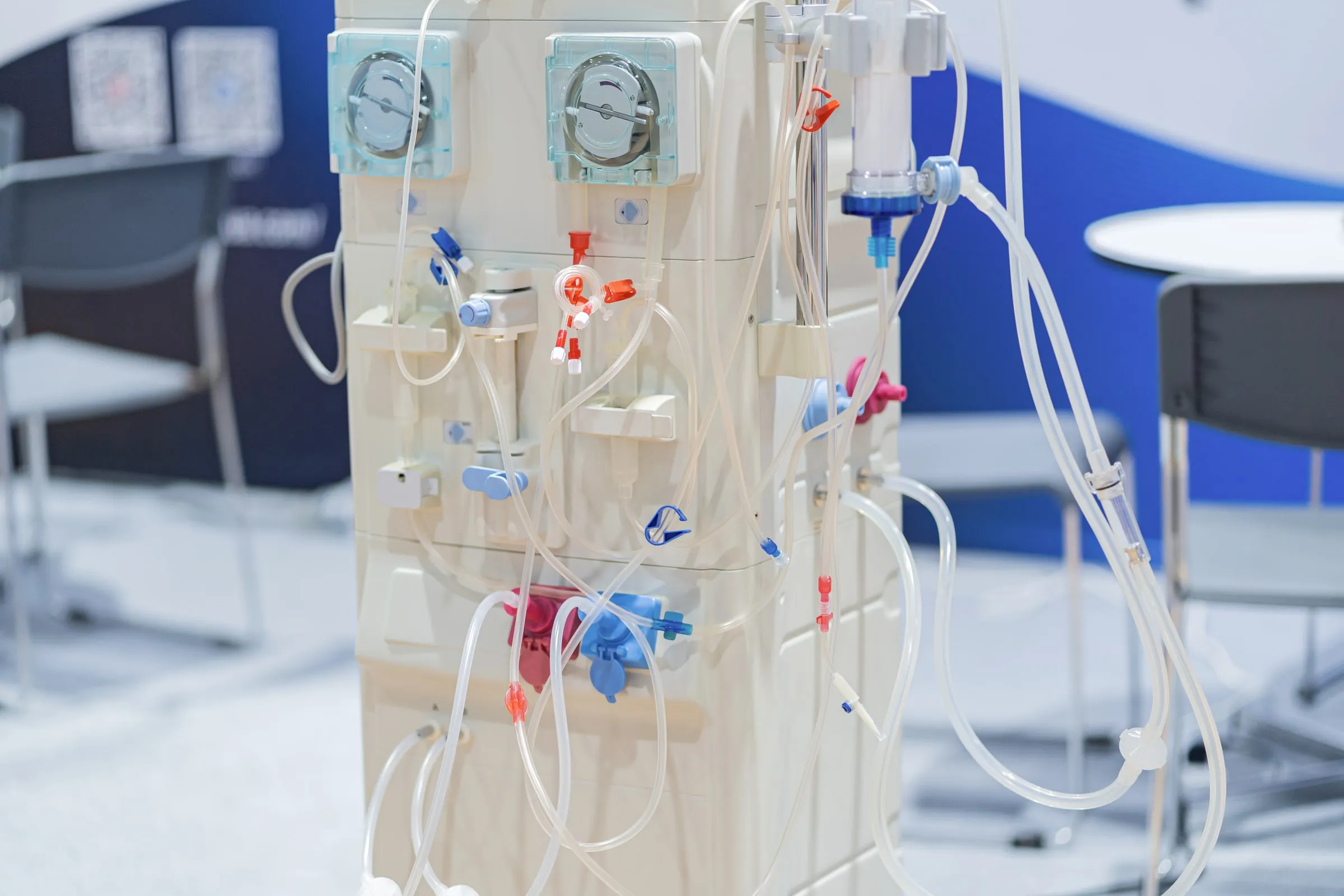 Close-up of a dialysis machine's tubing lines, drip chamber and control dials, with clinic chairs visible out of focus behind it