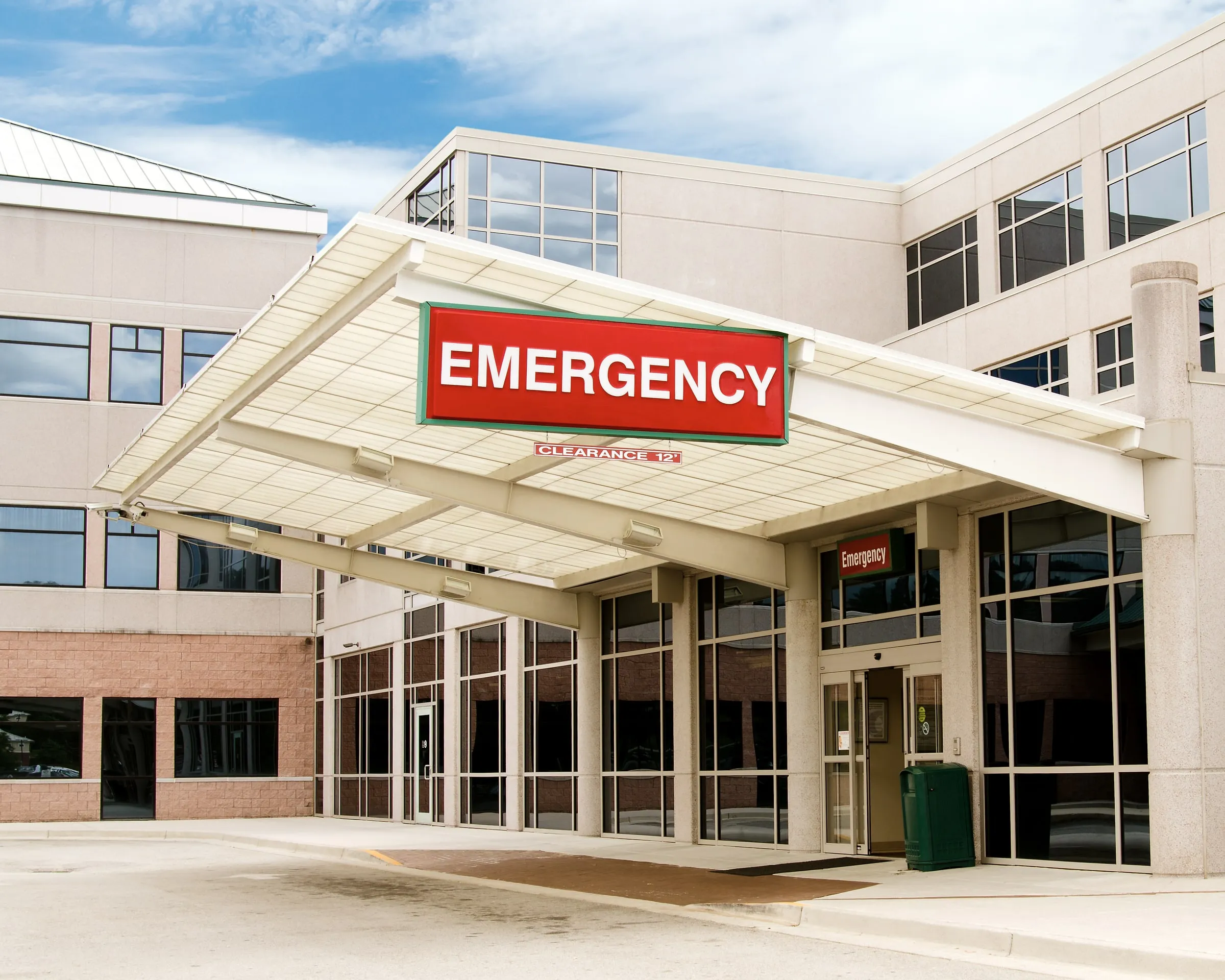 Illuminated red Emergency sign under a canopy at a hospital entrance, with a smaller Emergency sign beside the glass doors below