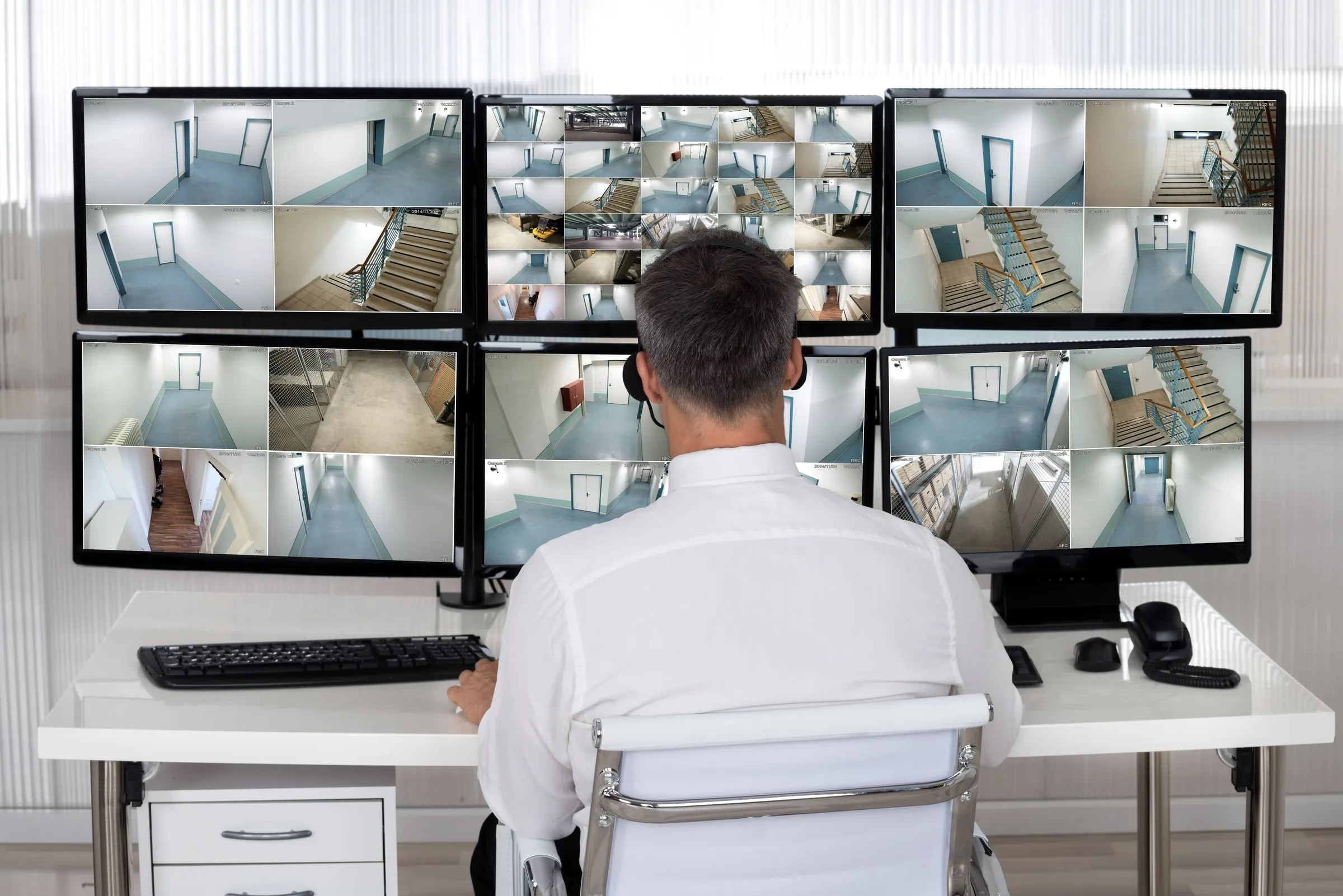 Officer seen from behind at a desk of six monitors showing hospital hallway and stairwell camera feeds, wearing a headset