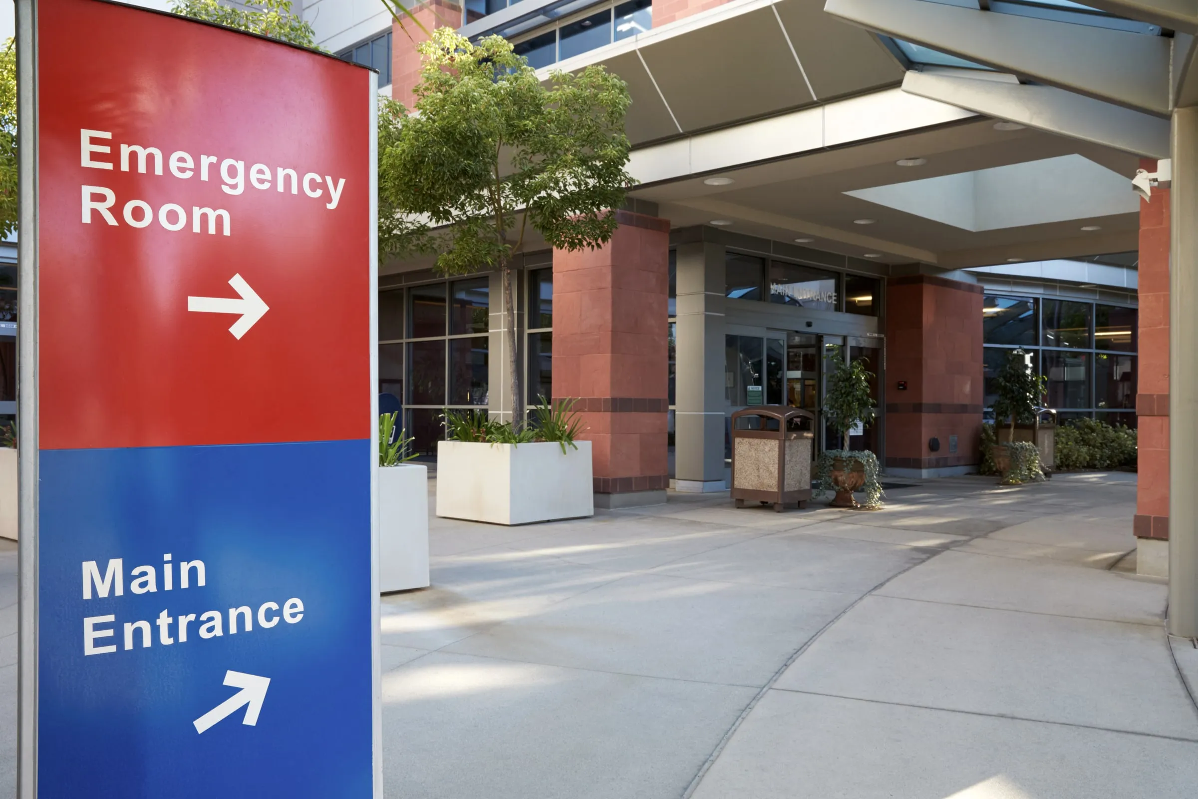 Red Emergency Room and blue Main Entrance directional signs mounted on a post outside a hospital building with a covered drop-off canopy
