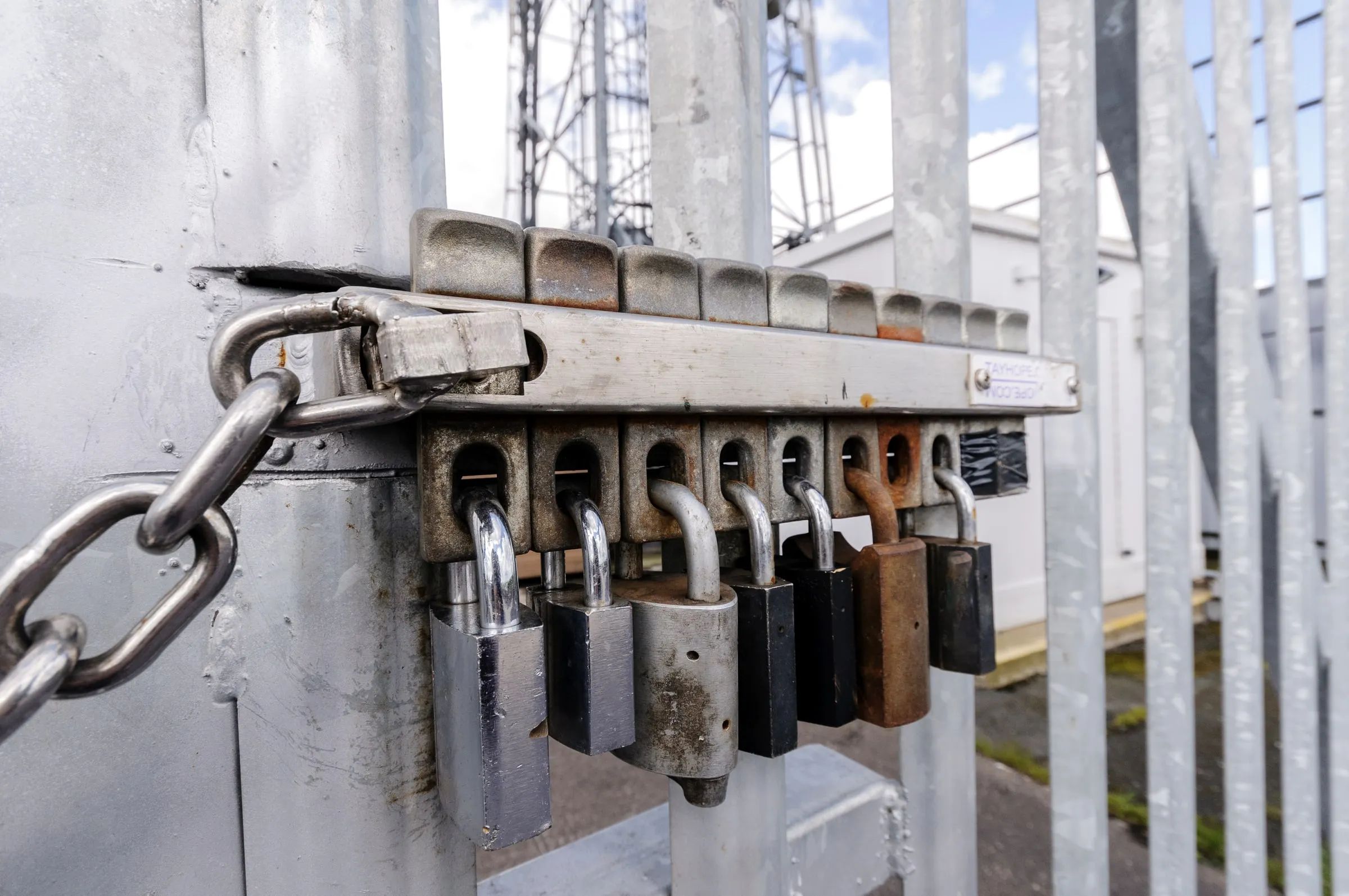 Chain-link gate secured with a length of chain and several padlocks of different sizes, with a lattice utility tower visible in the background