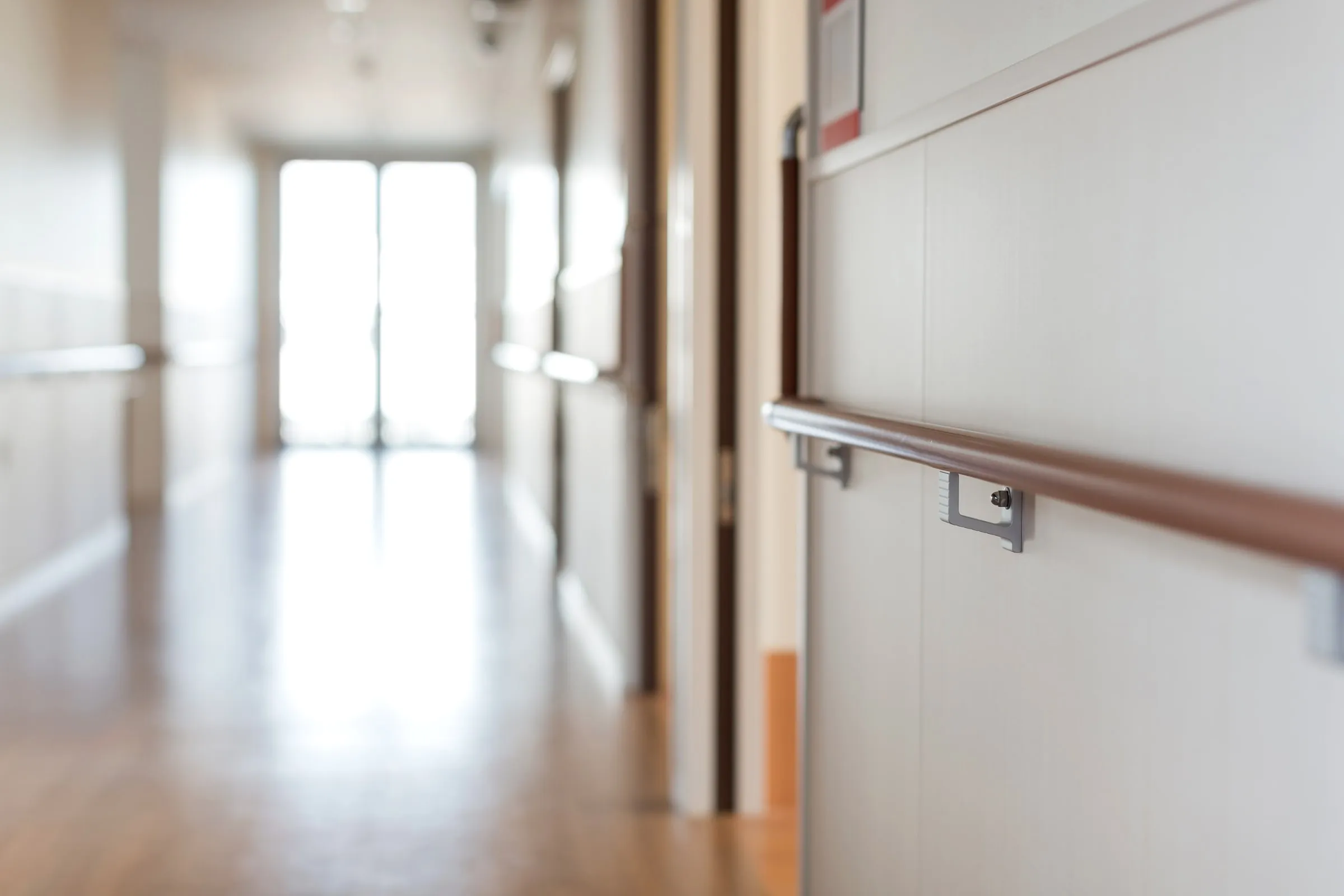 Interior hallway of a care facility with a wood-topped steel handrail mounted along the wall, doors and a sunlit exit at the far end