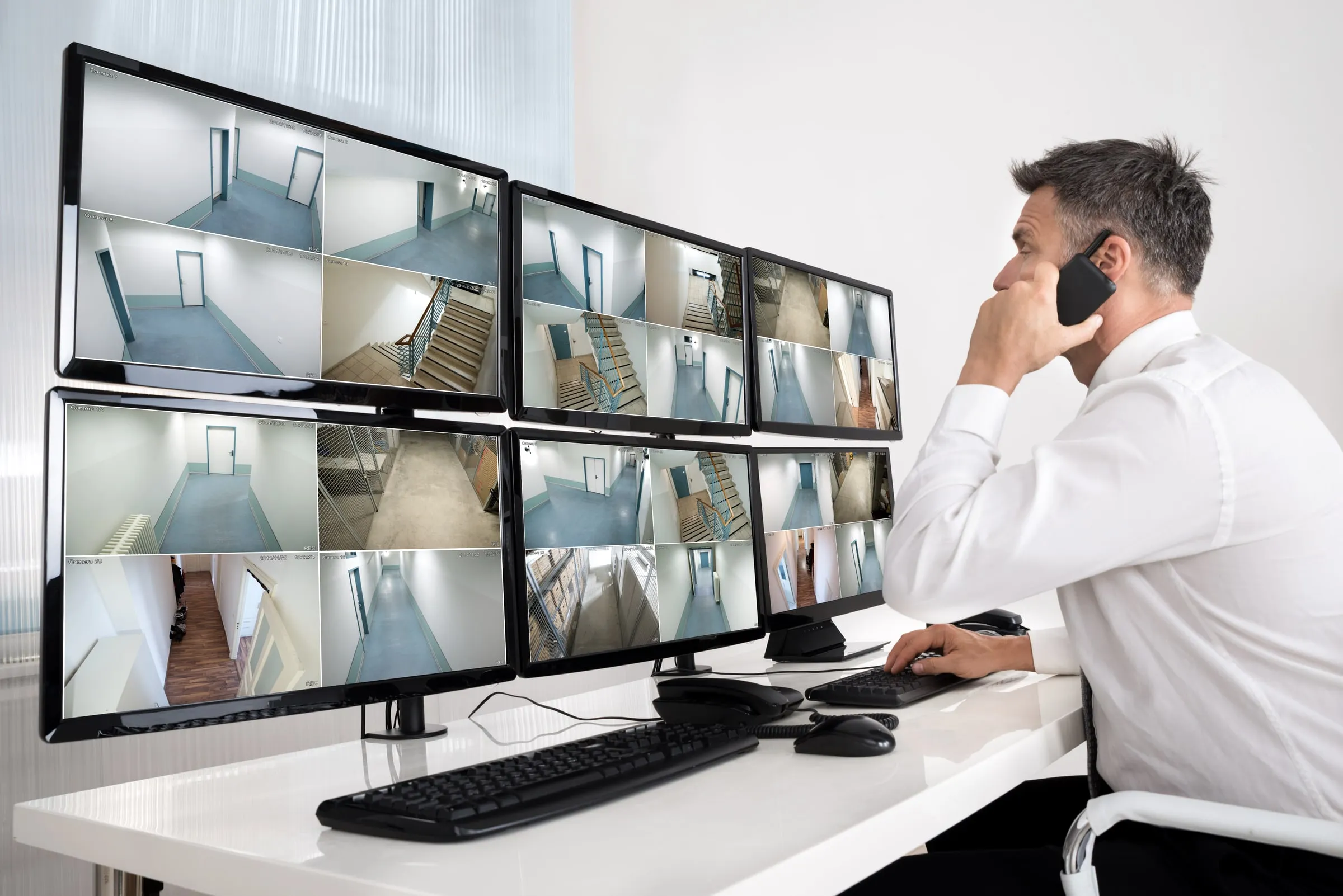 Man in a white shirt on the phone while looking at a bank of six monitors showing hallway and stairwell camera feeds at a desk