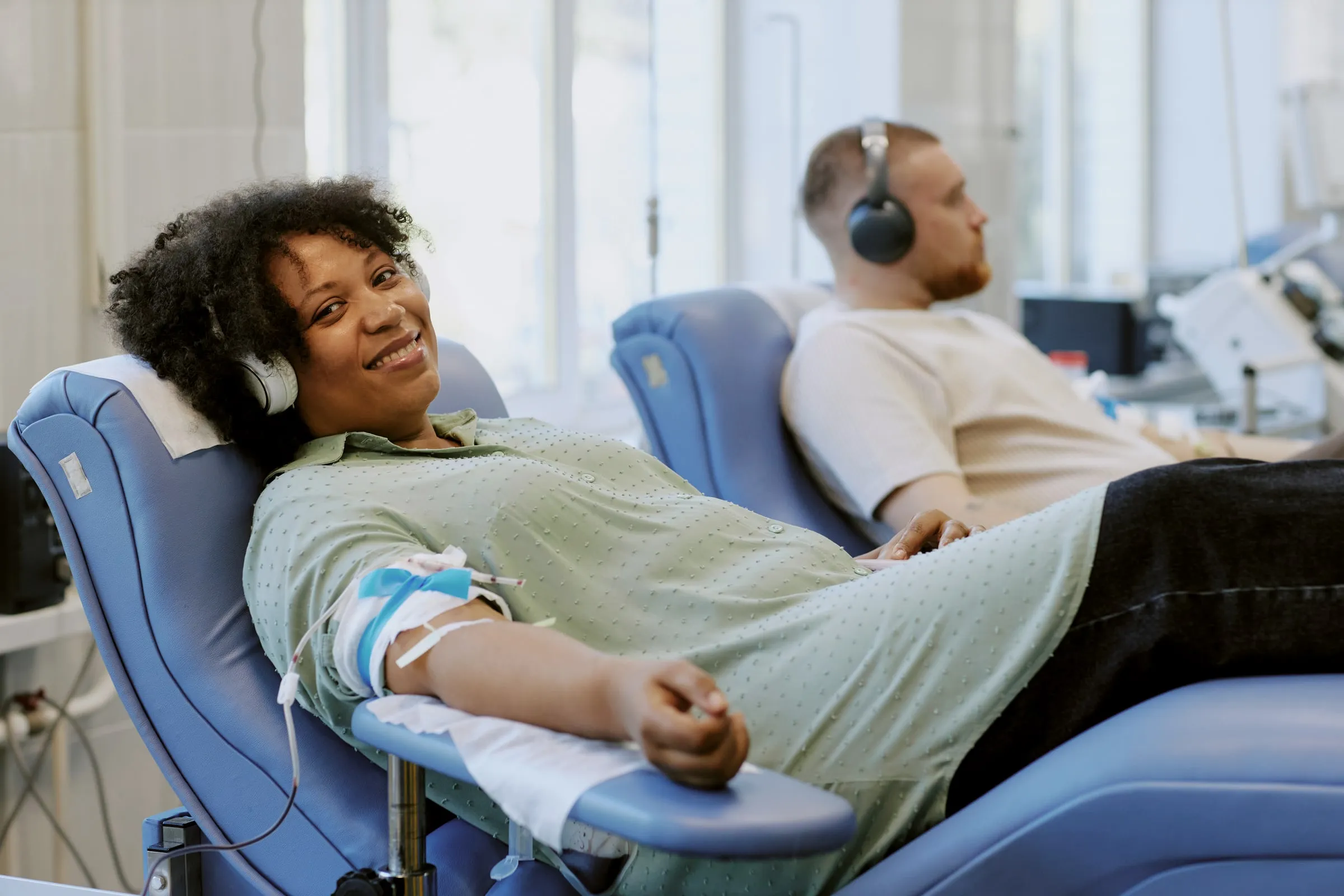 Two people reclined in blue donation chairs wearing headphones, one with an arm wrapped in gauze and tubing connected to donation equipment