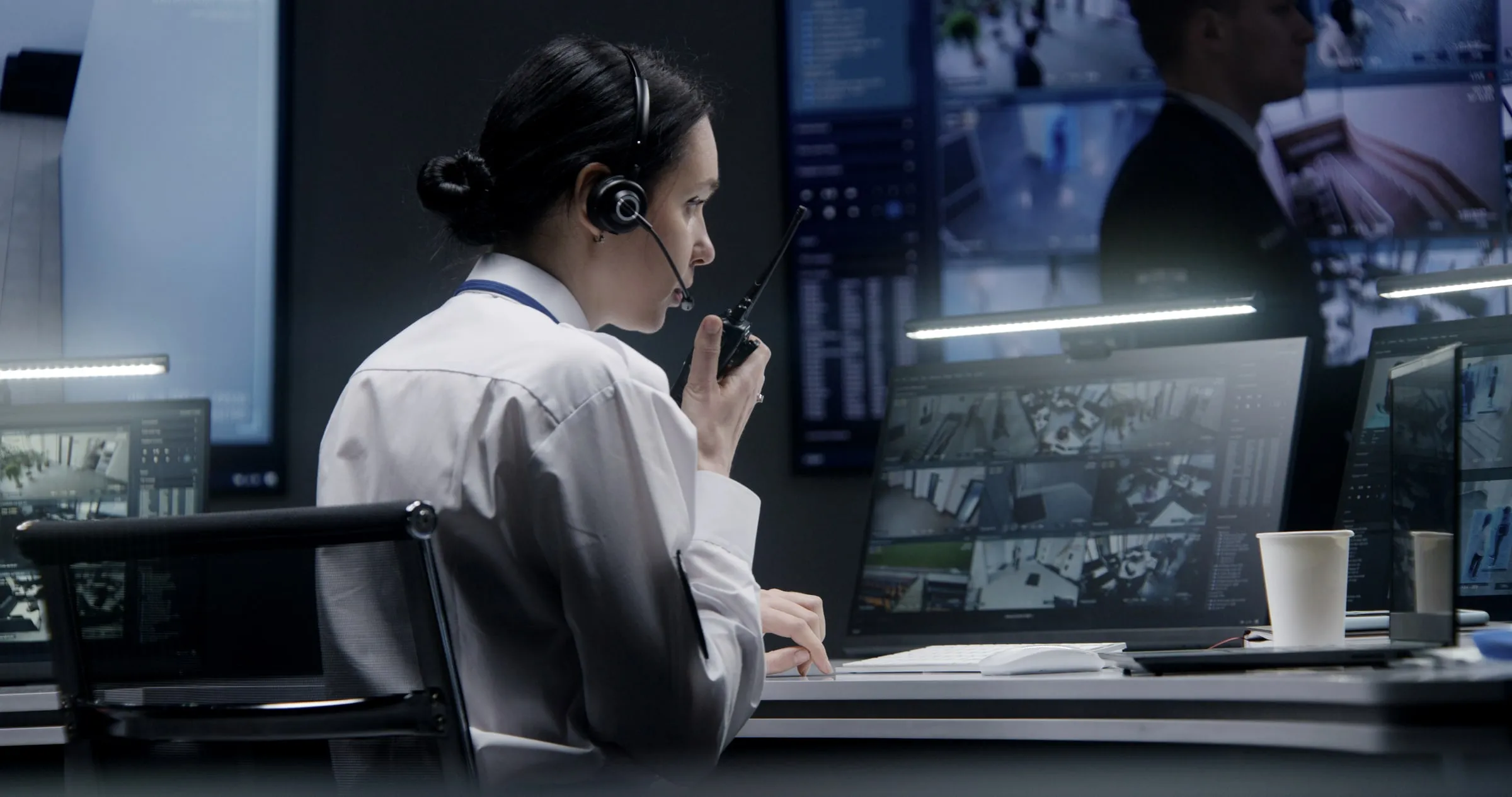 Security operator wearing a headset speaks into a handheld radio while facing a wall of monitor screens showing camera feeds, with coffee cups on the desk