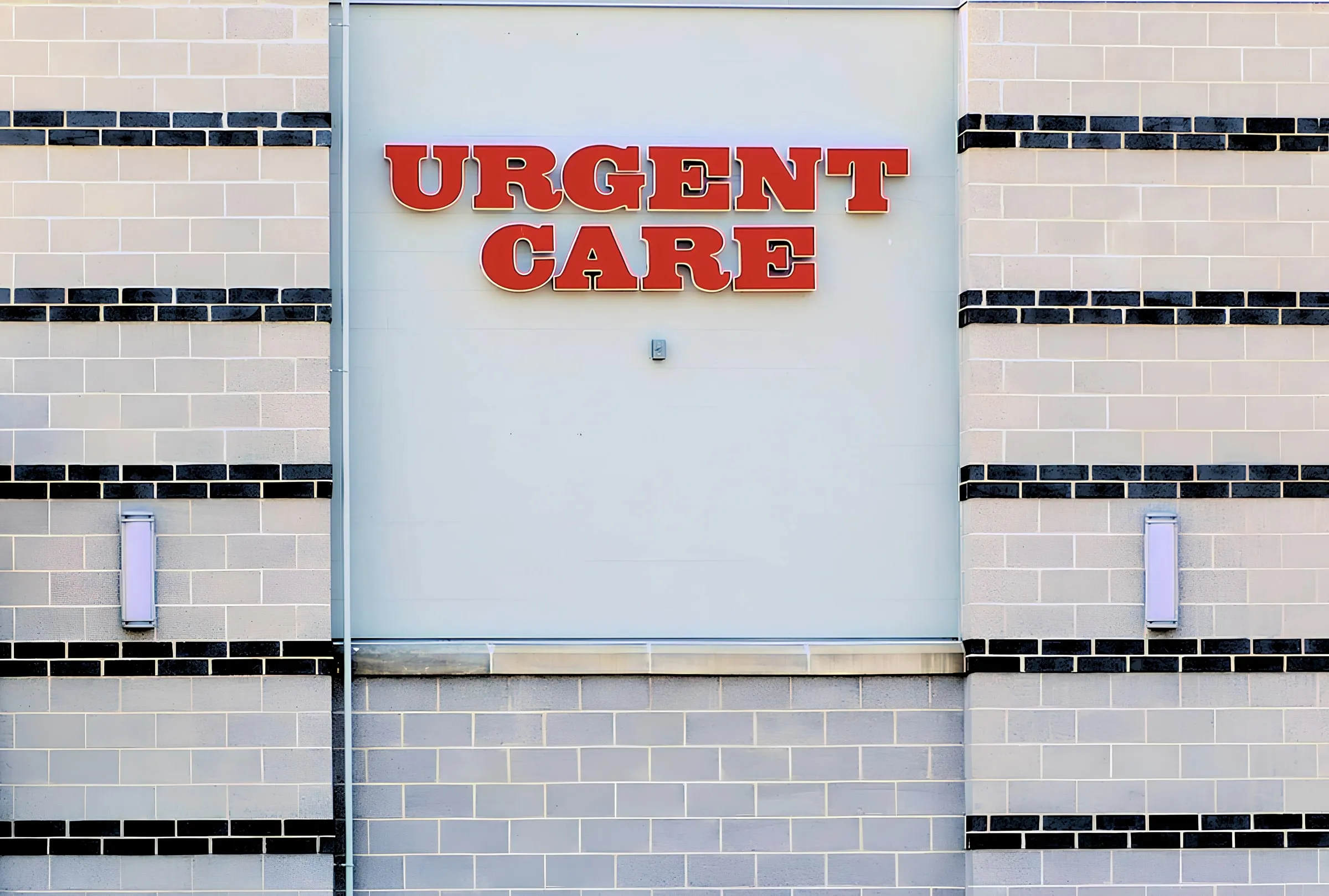 Red Urgent Care channel letters mounted on a gray and black tile building facade, above a plain signboard panel