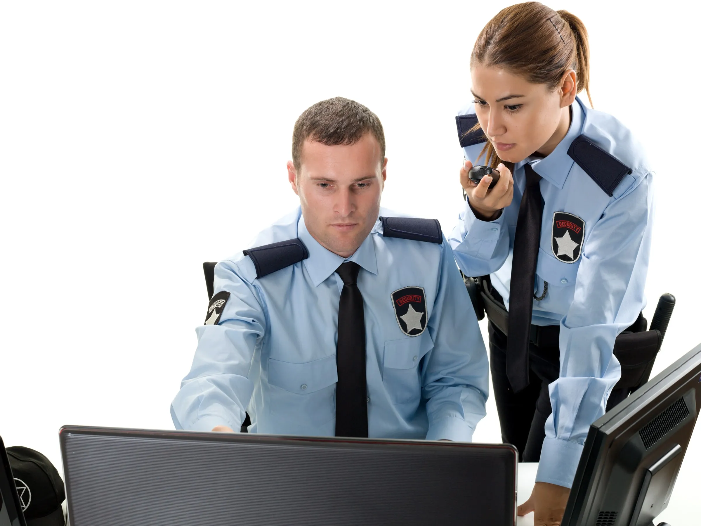 Two officers in light blue security shirts at a desk, one watching a monitor while the other speaks into a handheld radio