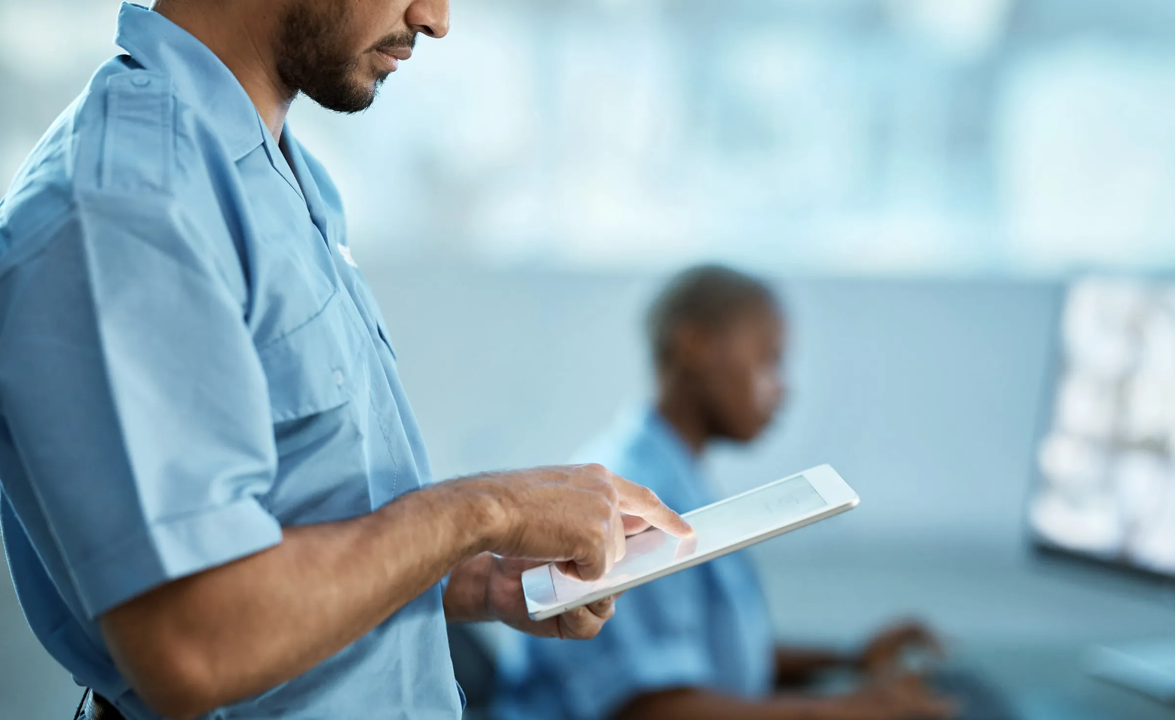 Security officer in a light blue shirt holding a tablet beside a colleague at a monitor desk