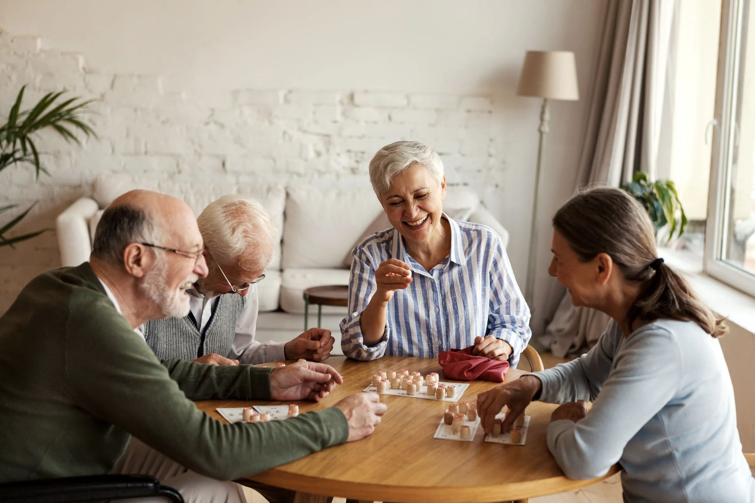 Four older adults laughing over a board game at a round wooden table in a bright living room