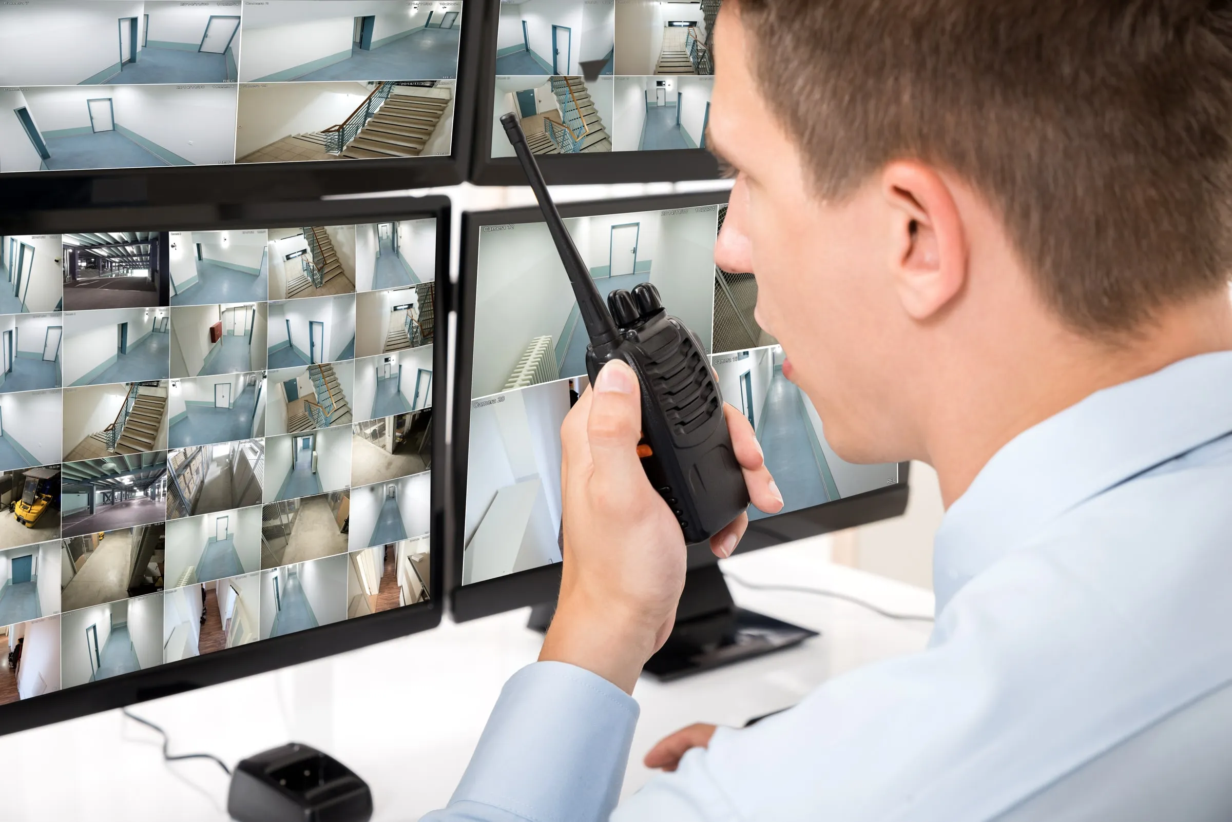 Officer seen from behind speaking into a handheld radio in front of monitors showing camera feeds of corridors, stairs and a garage