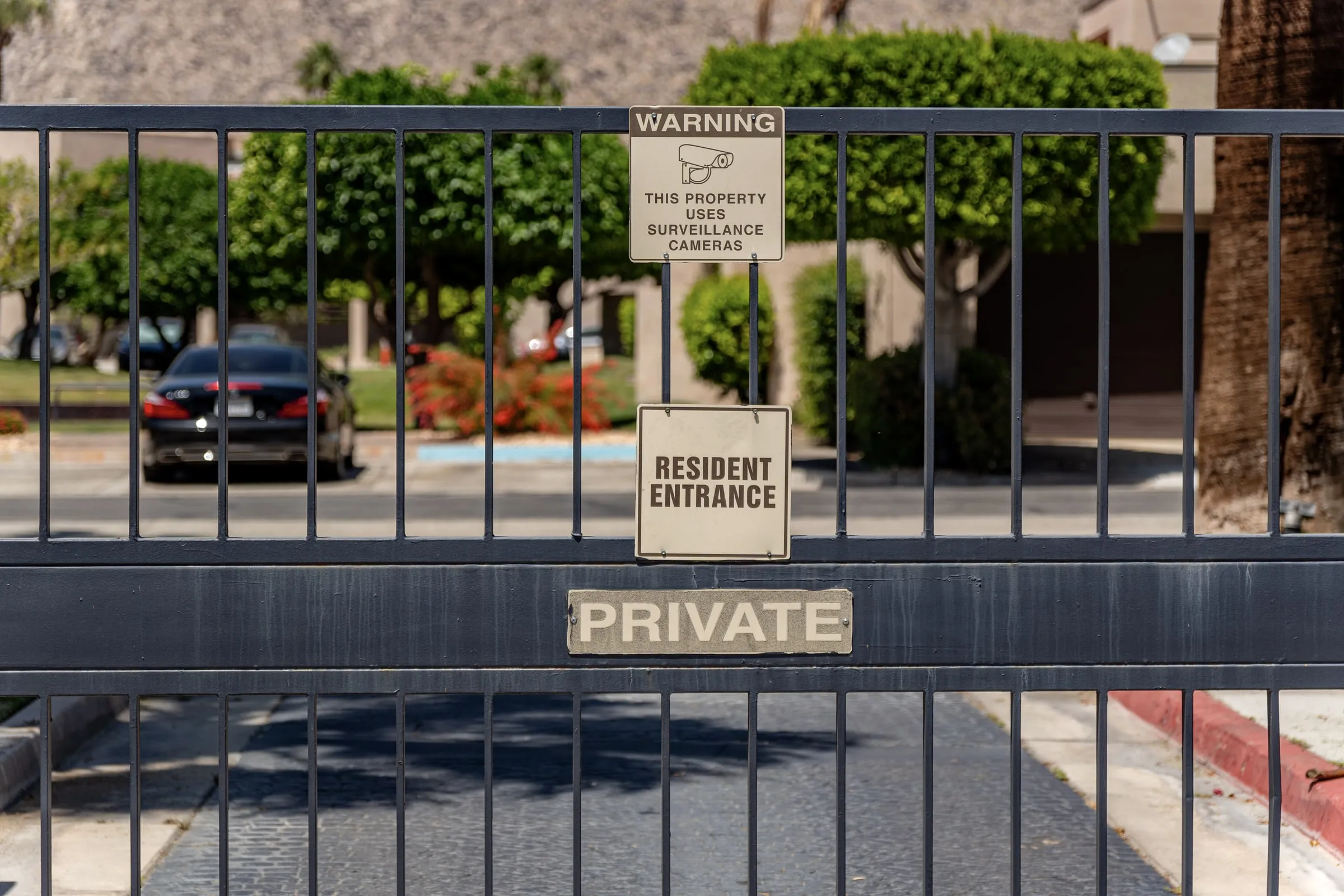 Dark metal entry gate carrying Private, Resident Entrance and surveillance camera warning signs, with parked cars and trees beyond