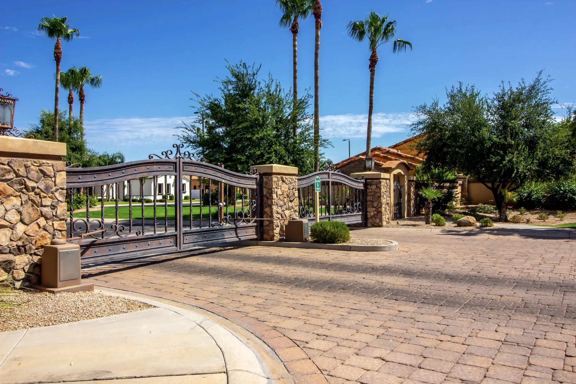 Ornate wrought-iron entry gates between stone pillars on a brick-paver drive, with palm trees and a tile-roofed gatehouse beyond