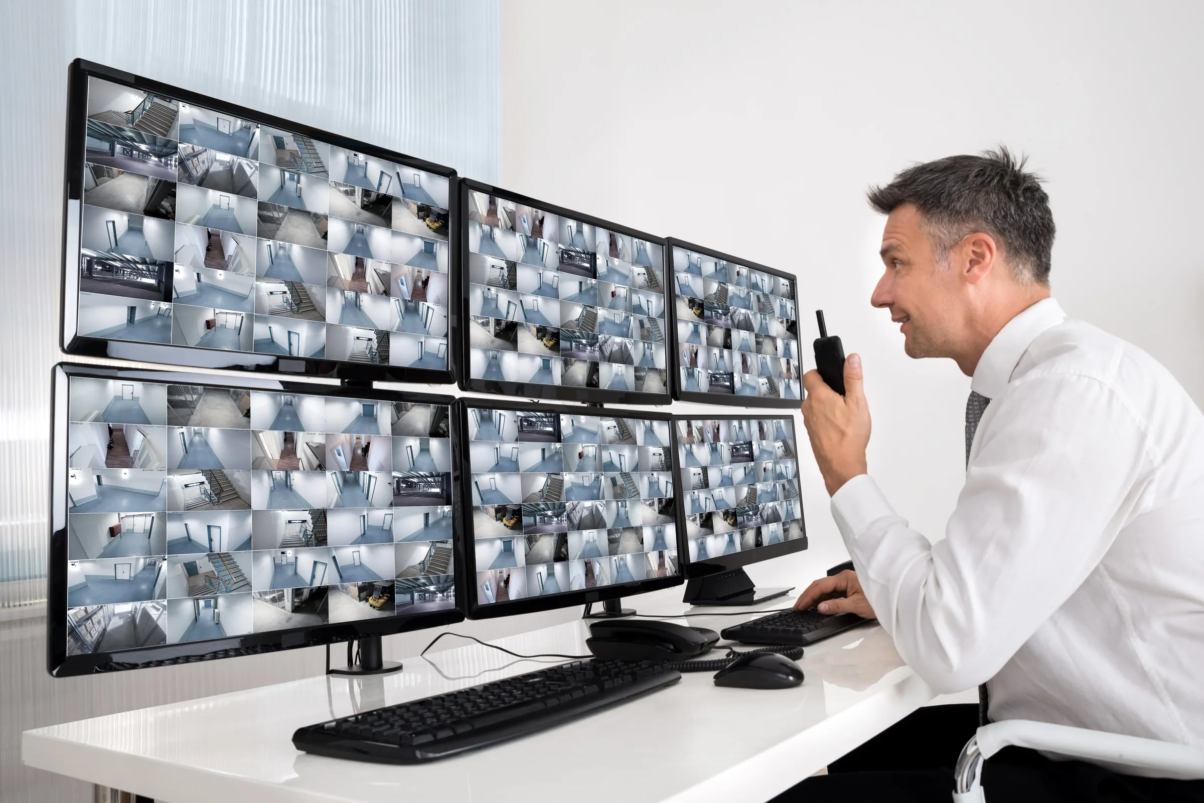 Man in a white shirt at a desk speaking into a radio in front of a bank of camera monitors