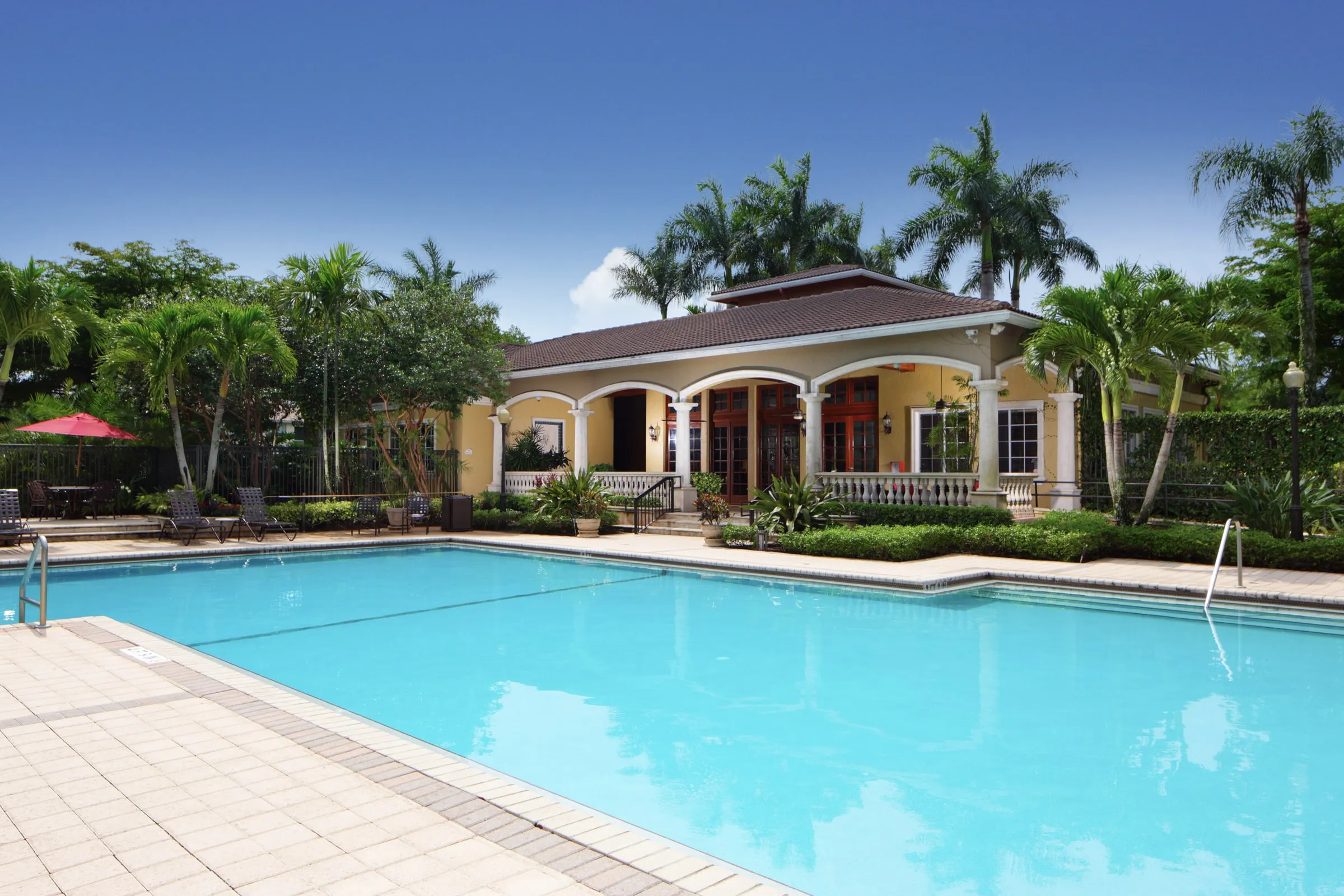 Community pool with clear blue water beside a yellow clubhouse with palms and a covered patio