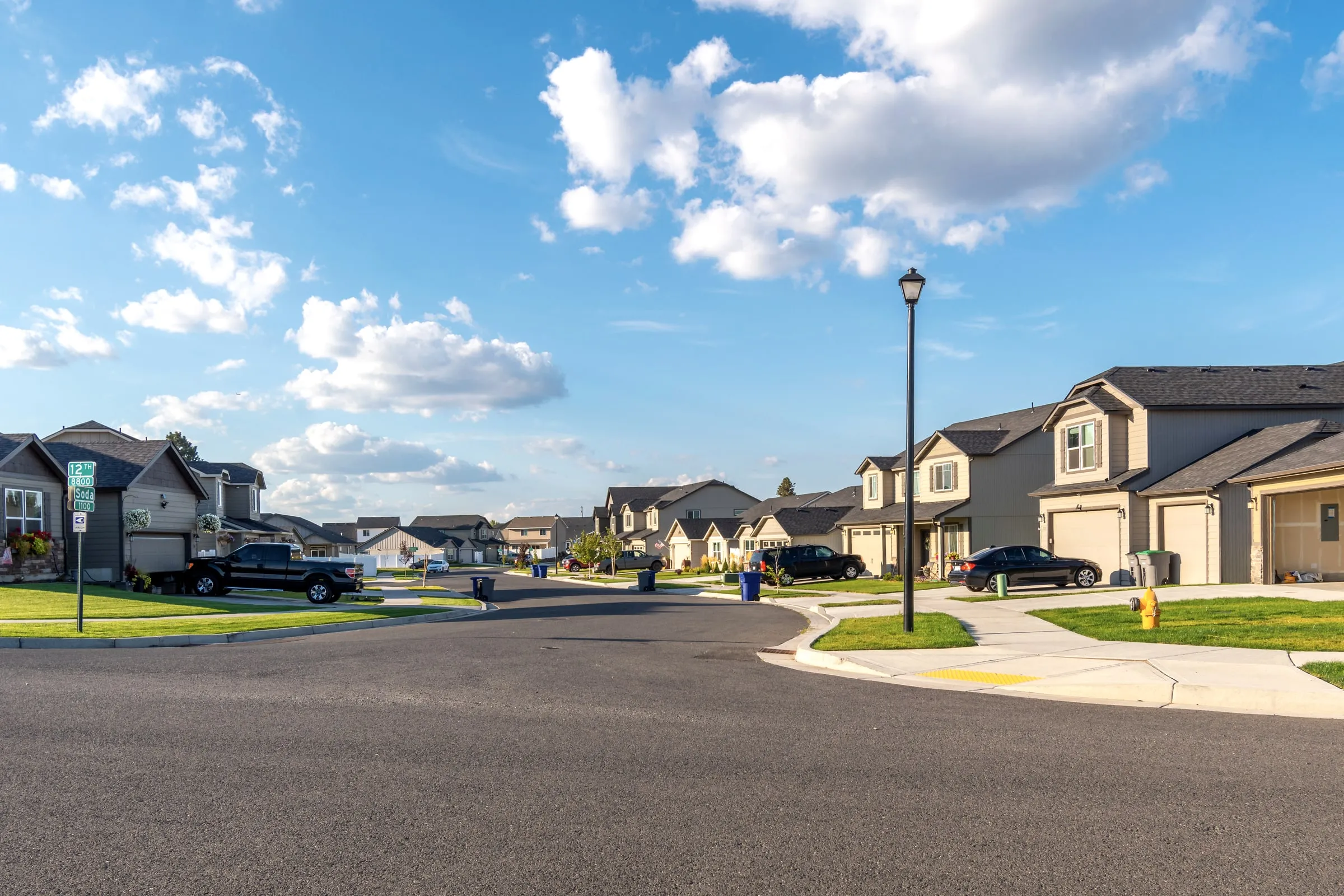 Suburban subdivision street with two-story houses, a parked black SUV and a landscaped corner under a blue sky