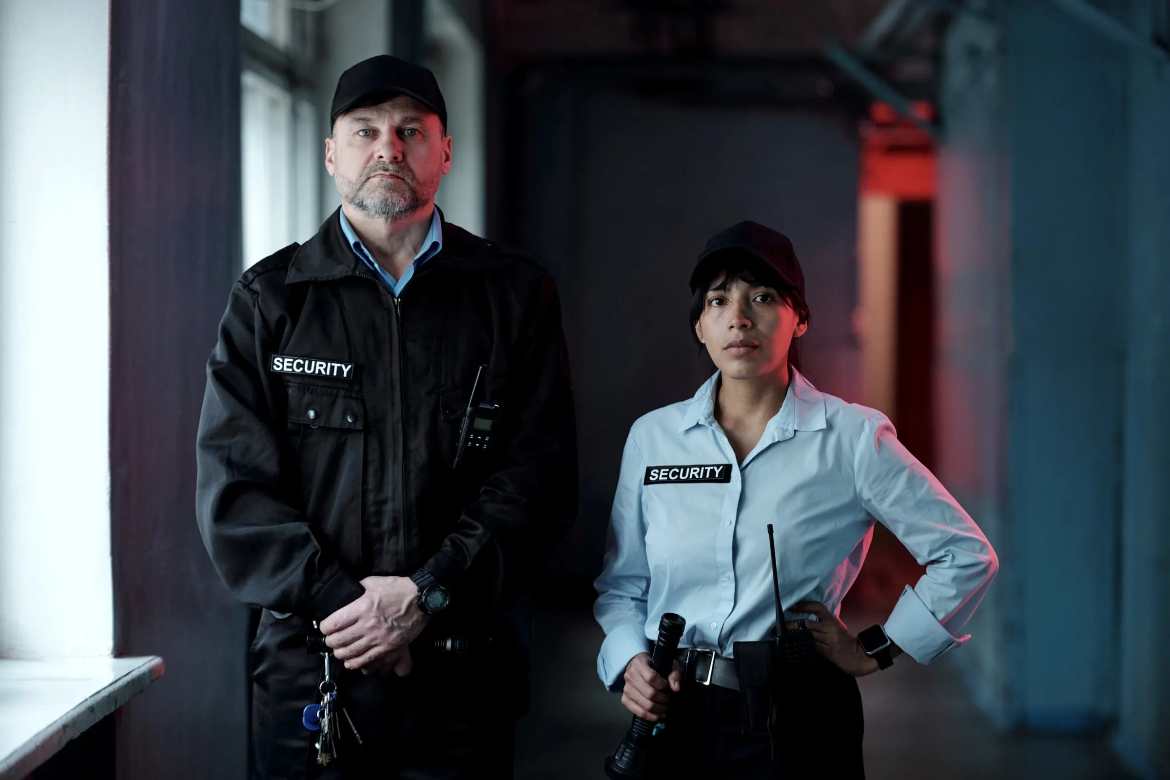 Two security officers in dark uniforms with radios standing at a doorway lit by a red light