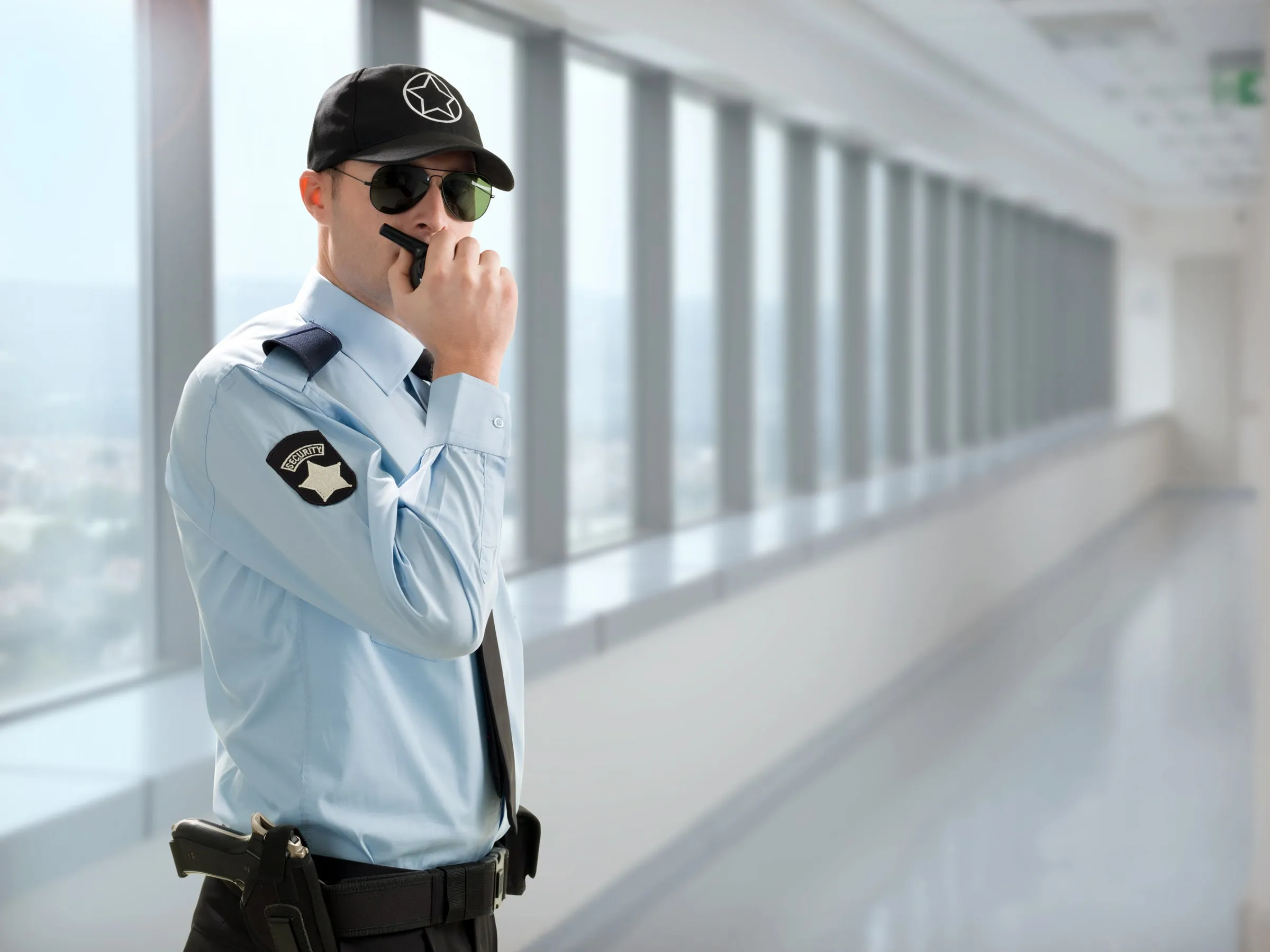 Security officer in a light uniform shirt and sunglasses speaking into a radio in a bright corridor