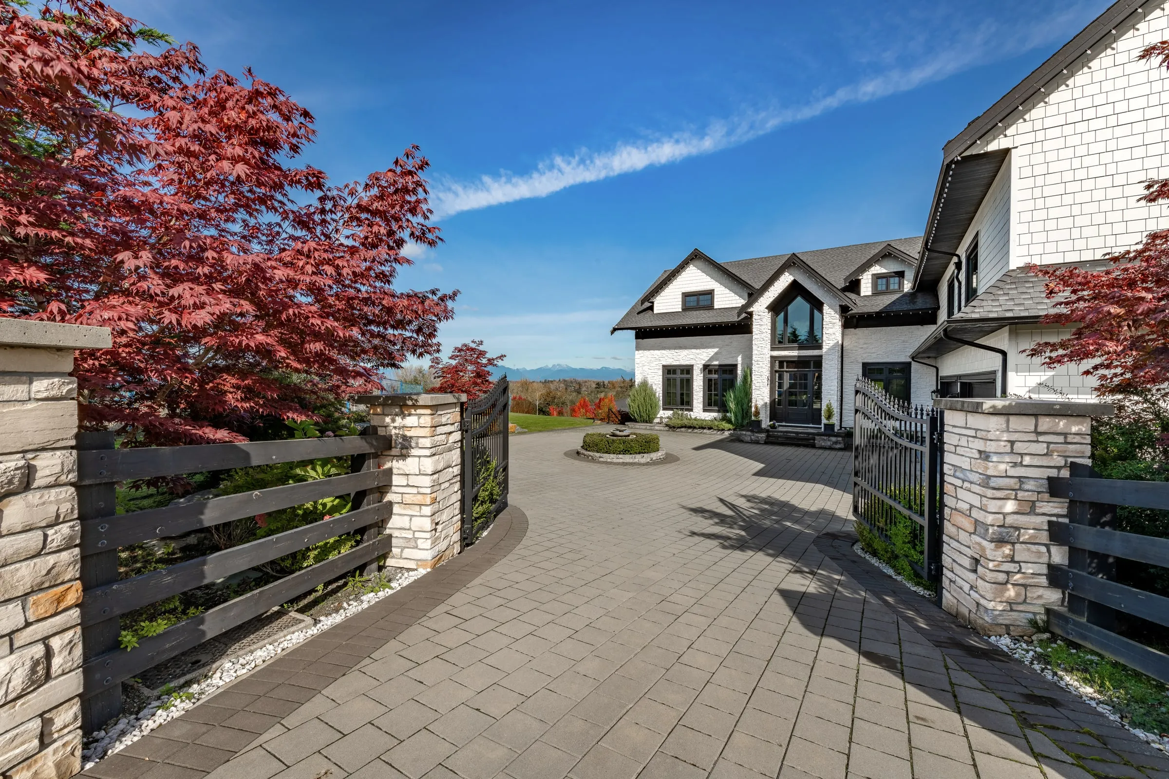Stone-and-iron entry gate opening onto a paved drive toward a large house with red maples