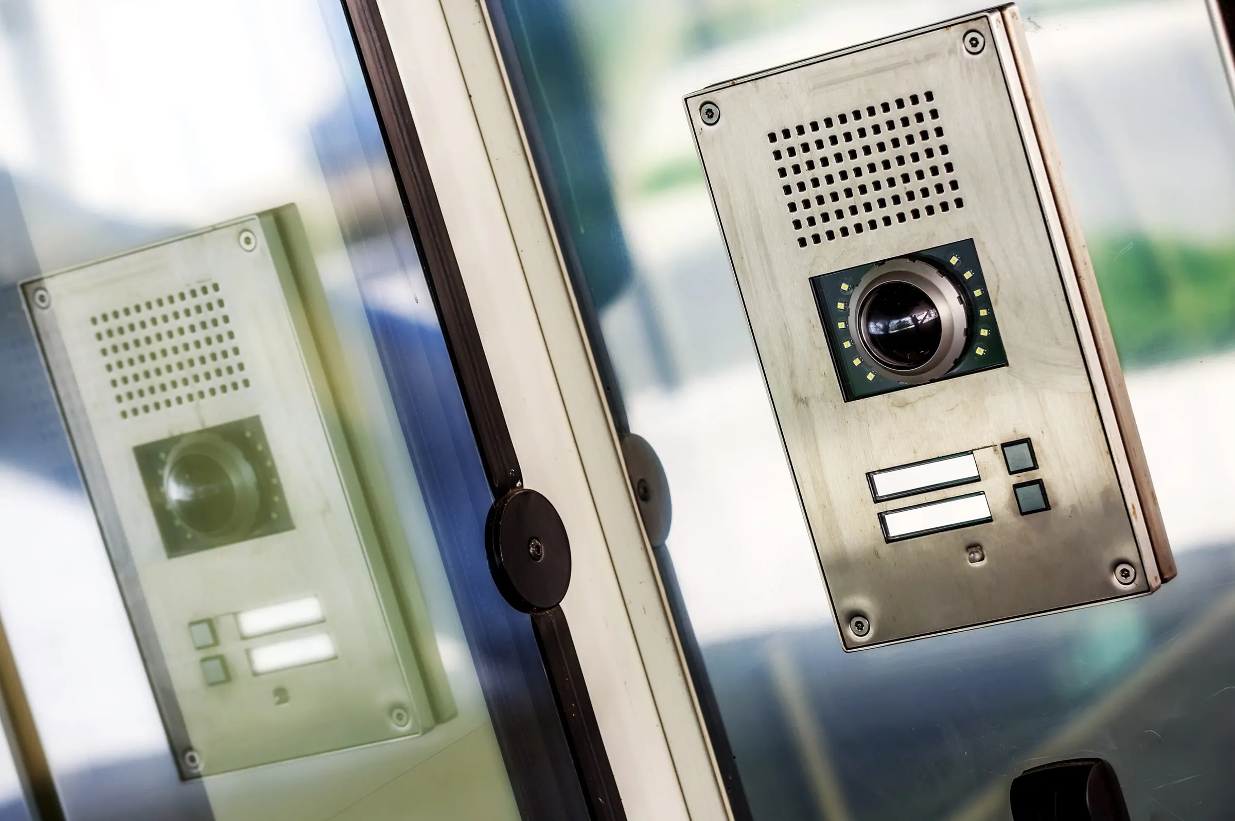 Stainless steel video intercom panel with a camera lens and call buttons mounted beside a glass entrance door