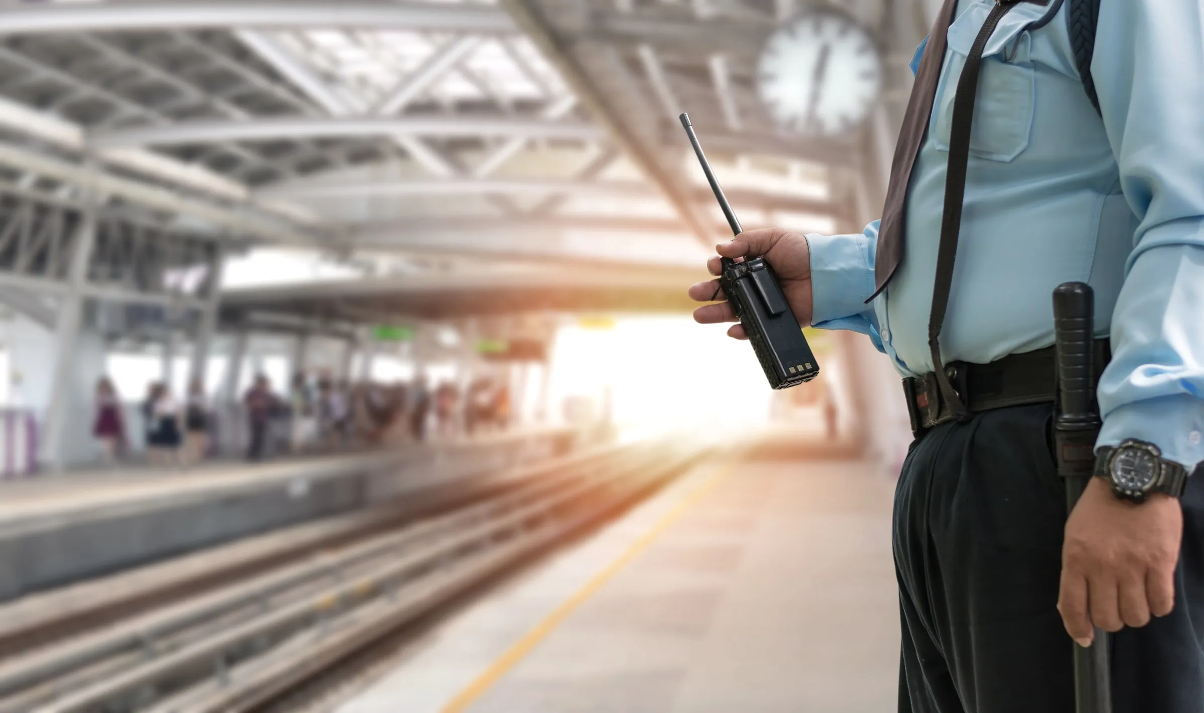 Security officer in a light blue shirt holding a handheld radio on a covered platform, with a wall clock overhead and a blurred crowd waiting beyond