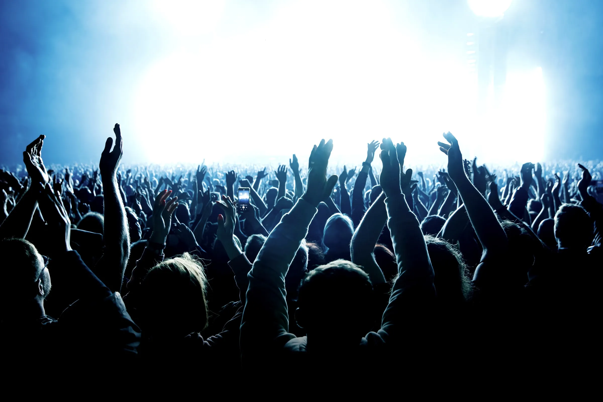 Crowd at a concert with raised hands silhouetted against bright blue stage lights