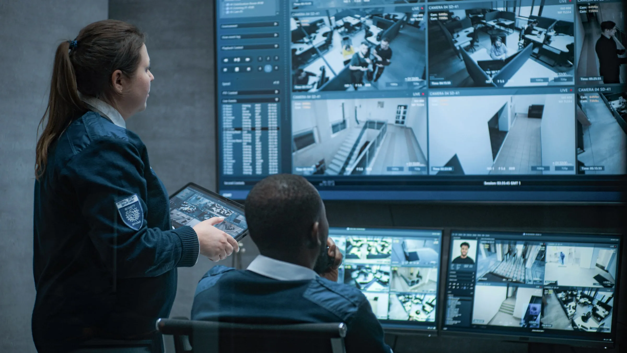 Security officer in a dark uniform pointing at a bank of camera monitors beside a colleague