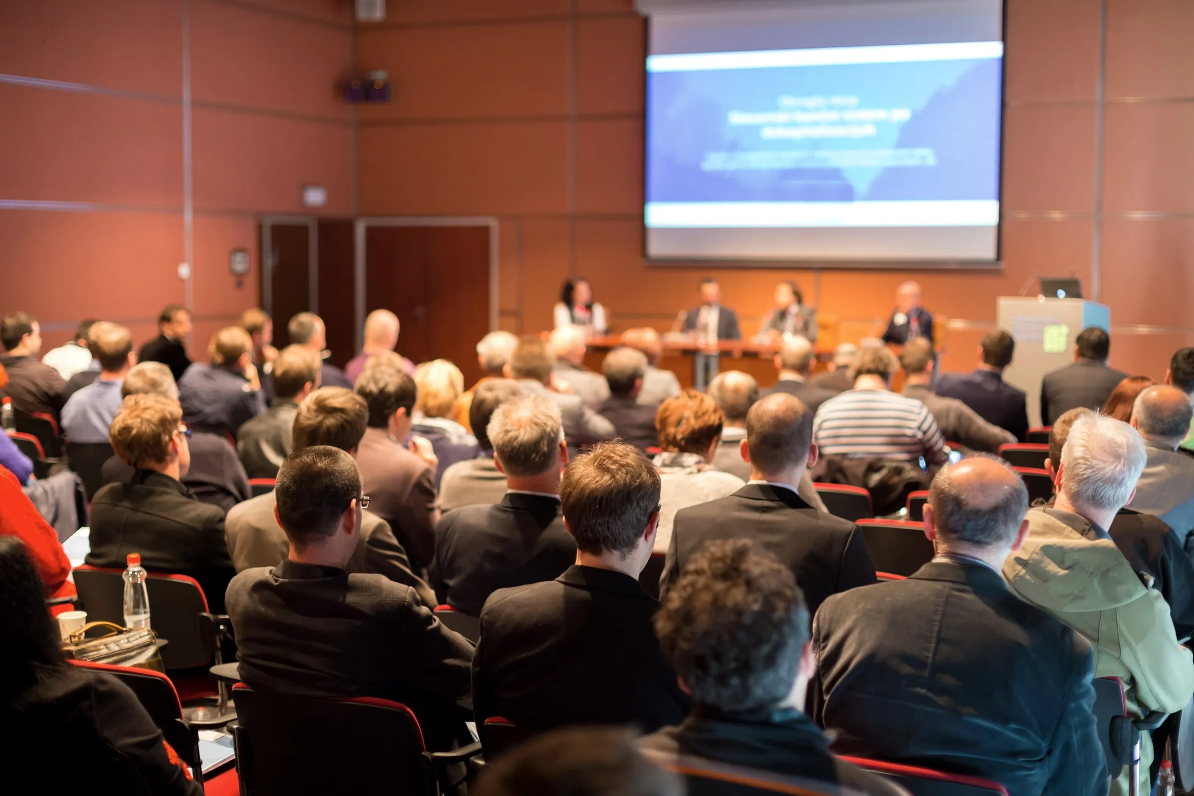 Audience seated in rows facing a panel and a projection screen at a conference