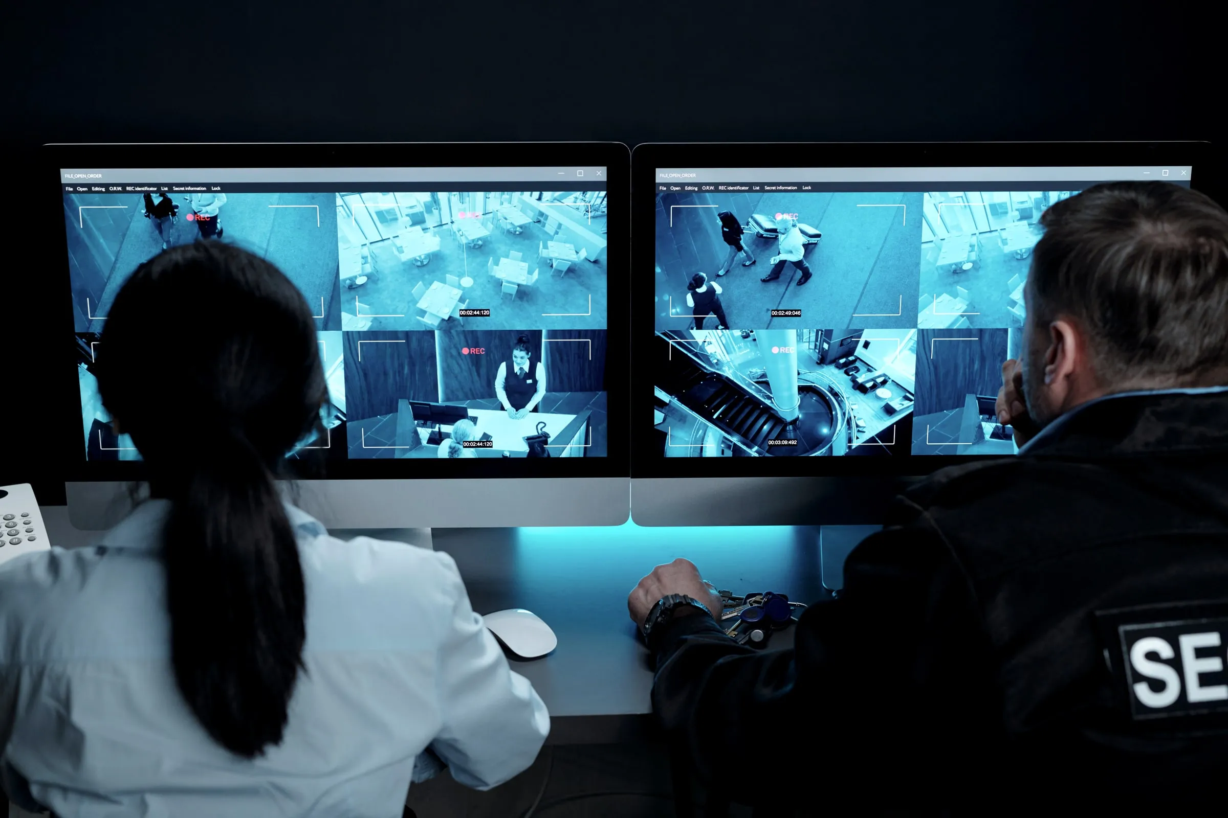 Two people seen from behind at a desk watching camera feeds of a lobby and a dining area on two monitors, one wearing a jacket marked SECURITY