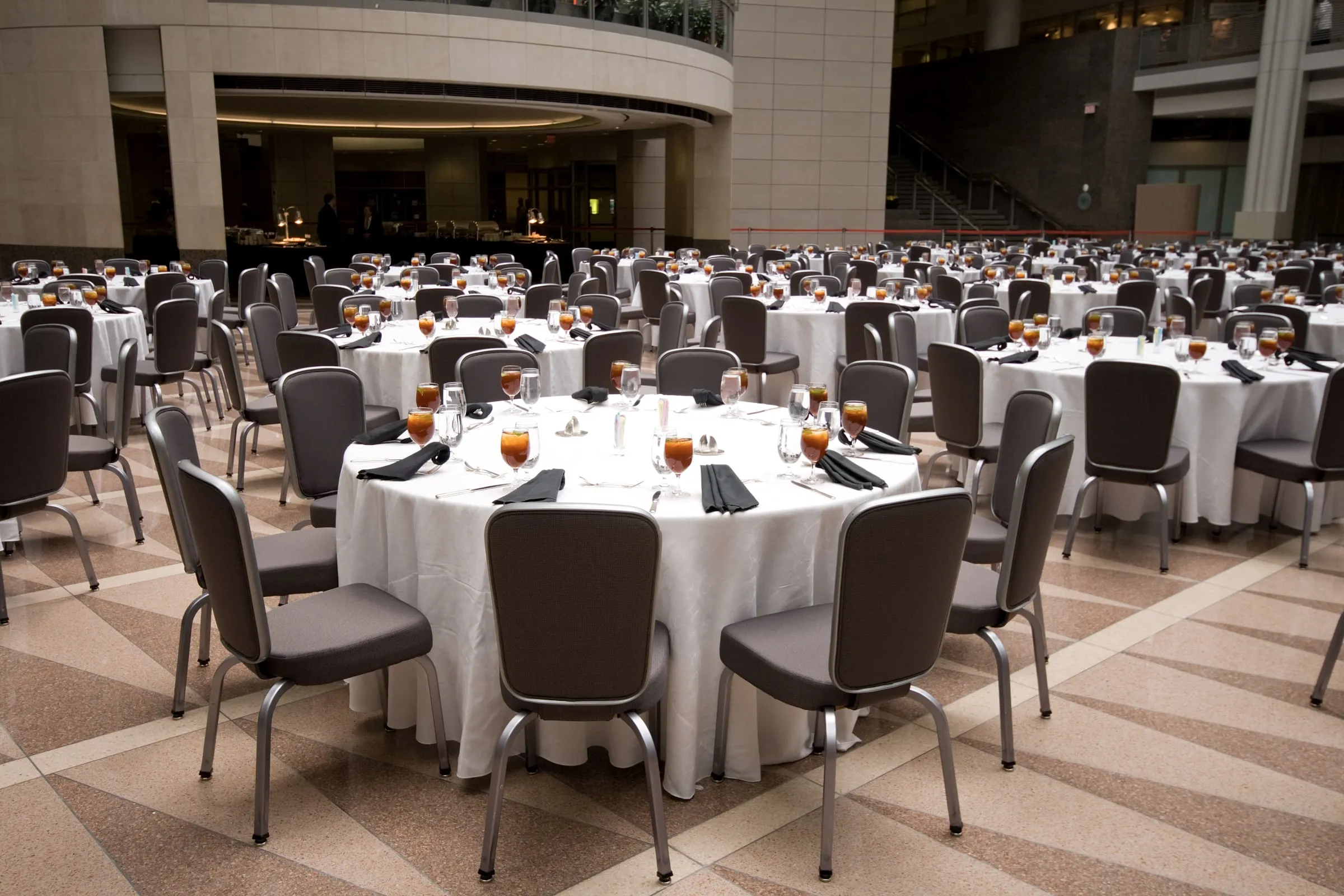 Banquet hall set with round tables in white linens, dark upholstered chairs and iced tea glasses, with a buffet line and a mezzanine at the back