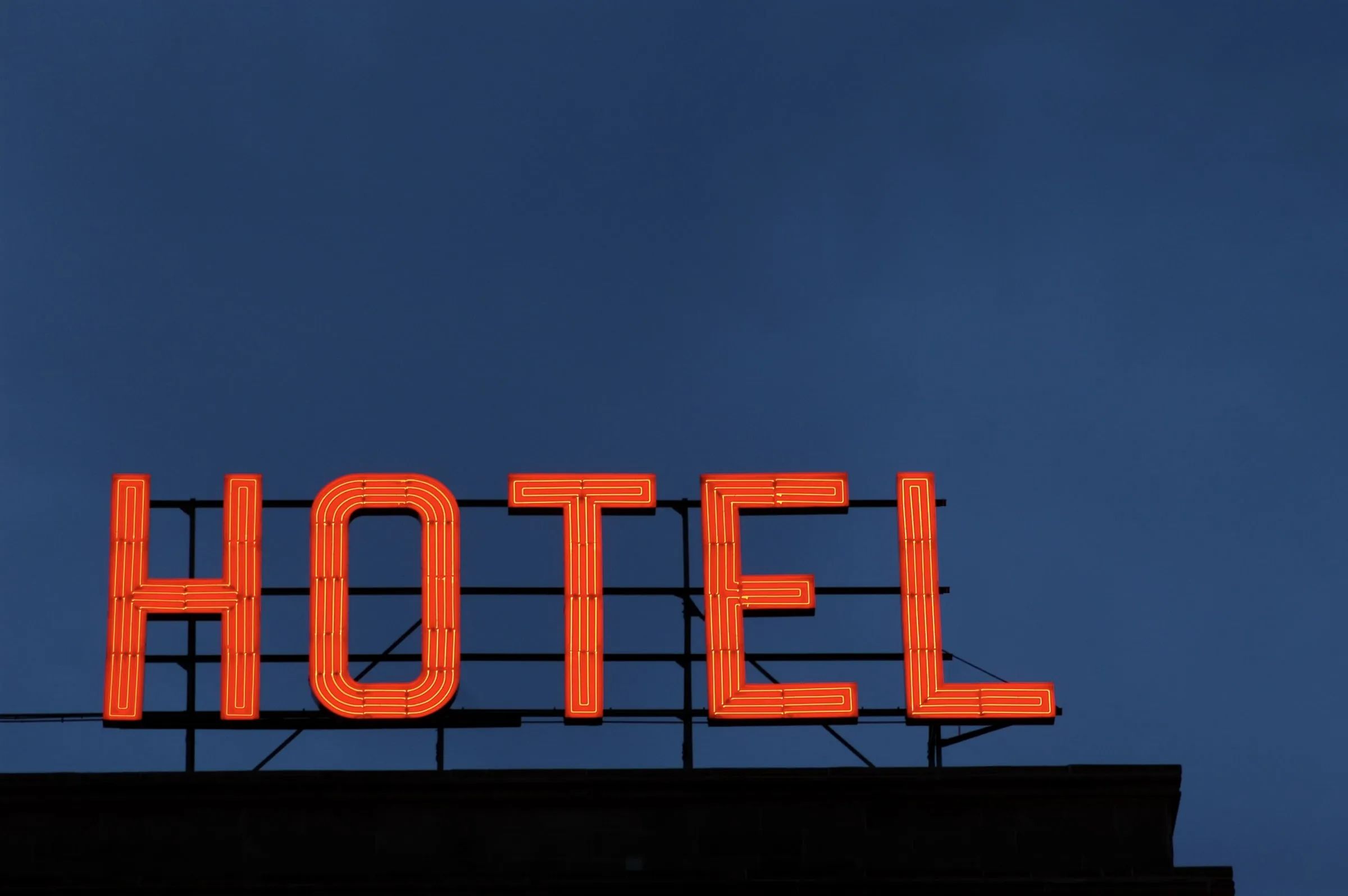 Red neon HOTEL sign on a rooftop steel frame against a deep blue evening sky